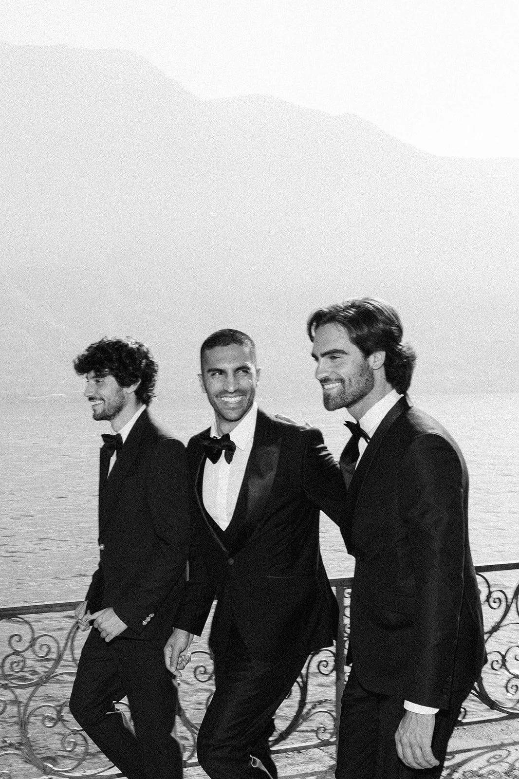 Three men in tuxedos standing outdoors, smiling, with water and mountains in the background.
