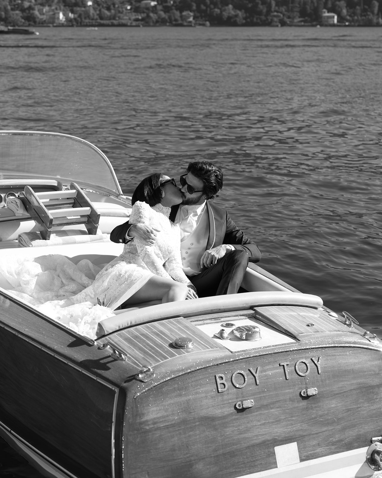 Black-and-white portrait of a bride and groom seated on a boat with the Lake Como mountains behind them.