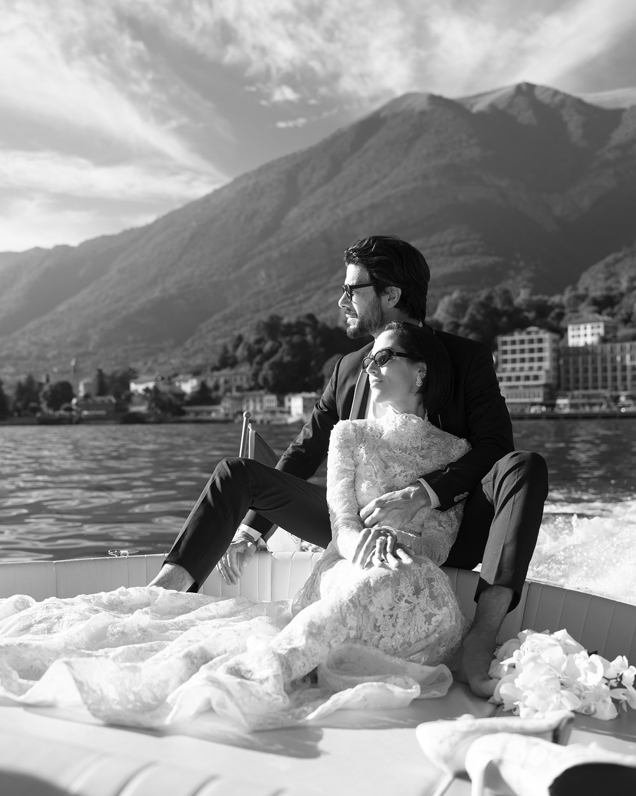 Black-and-white portrait of a bride and groom seated on a boat with the Lake Como mountains behind them.
