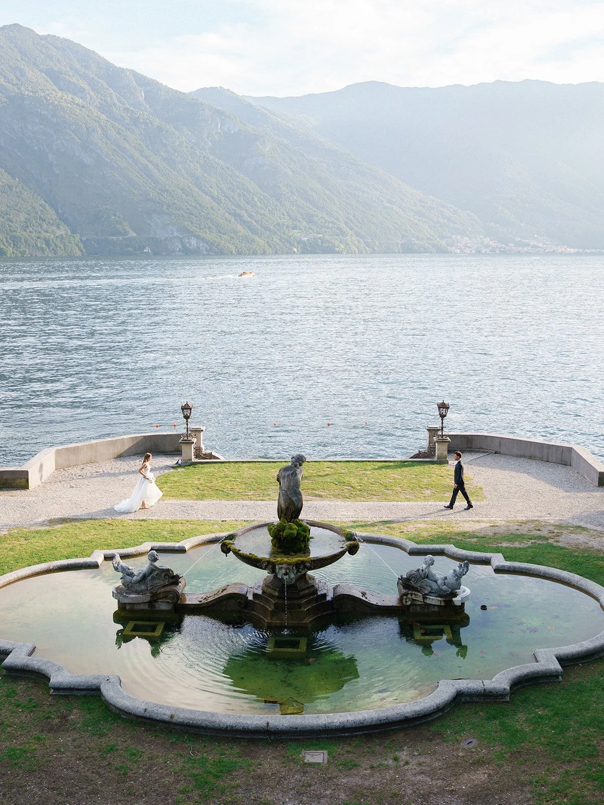 Historic fountain and formal lakeside gardens overlooking Lake Como, with a couple walking near the water.