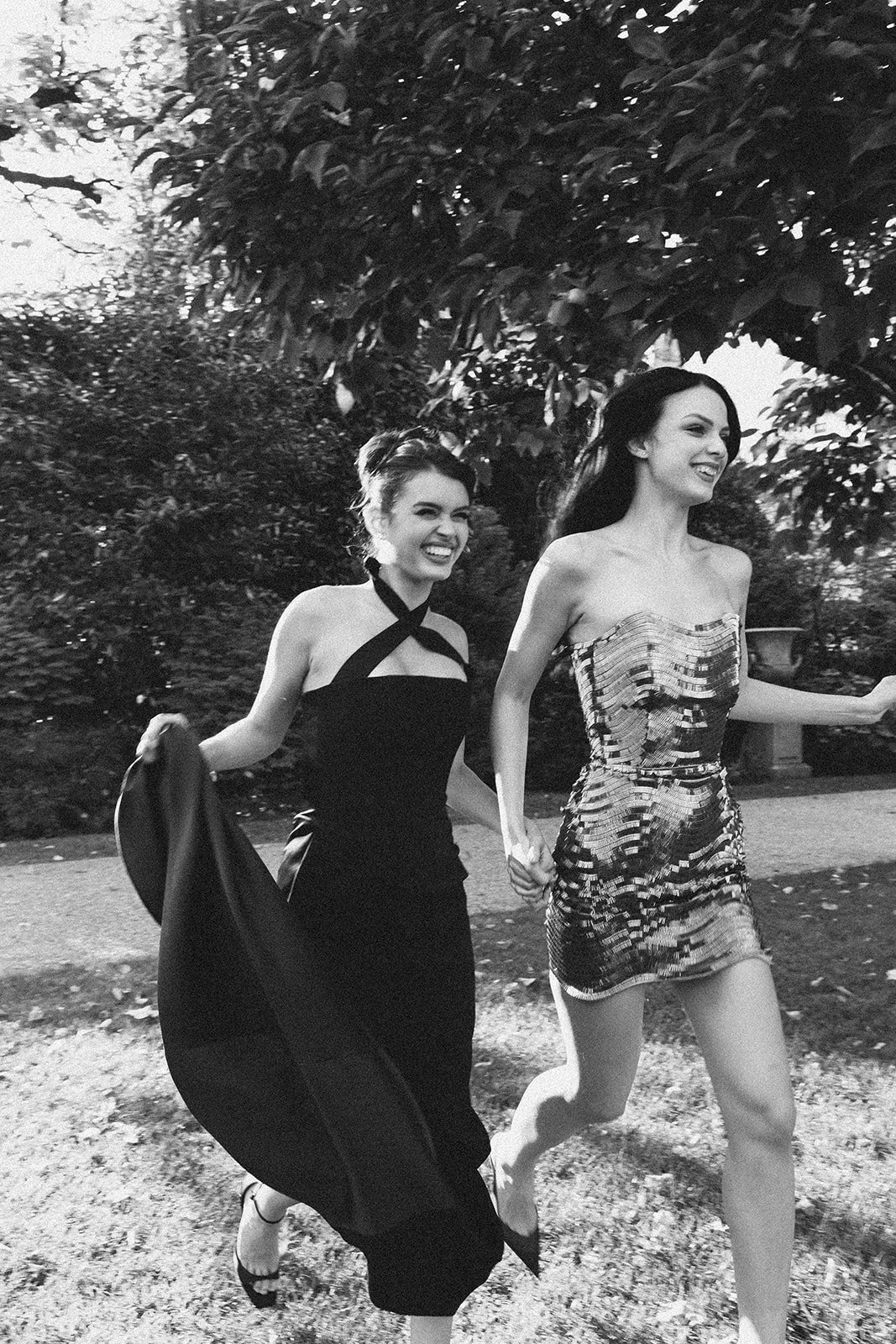 Two women walking outdoors in a park, smiling and holding hands. One is wearing a strapless, shiny dress, and the other is wearing a black dress with halter straps. The image is in black and white.