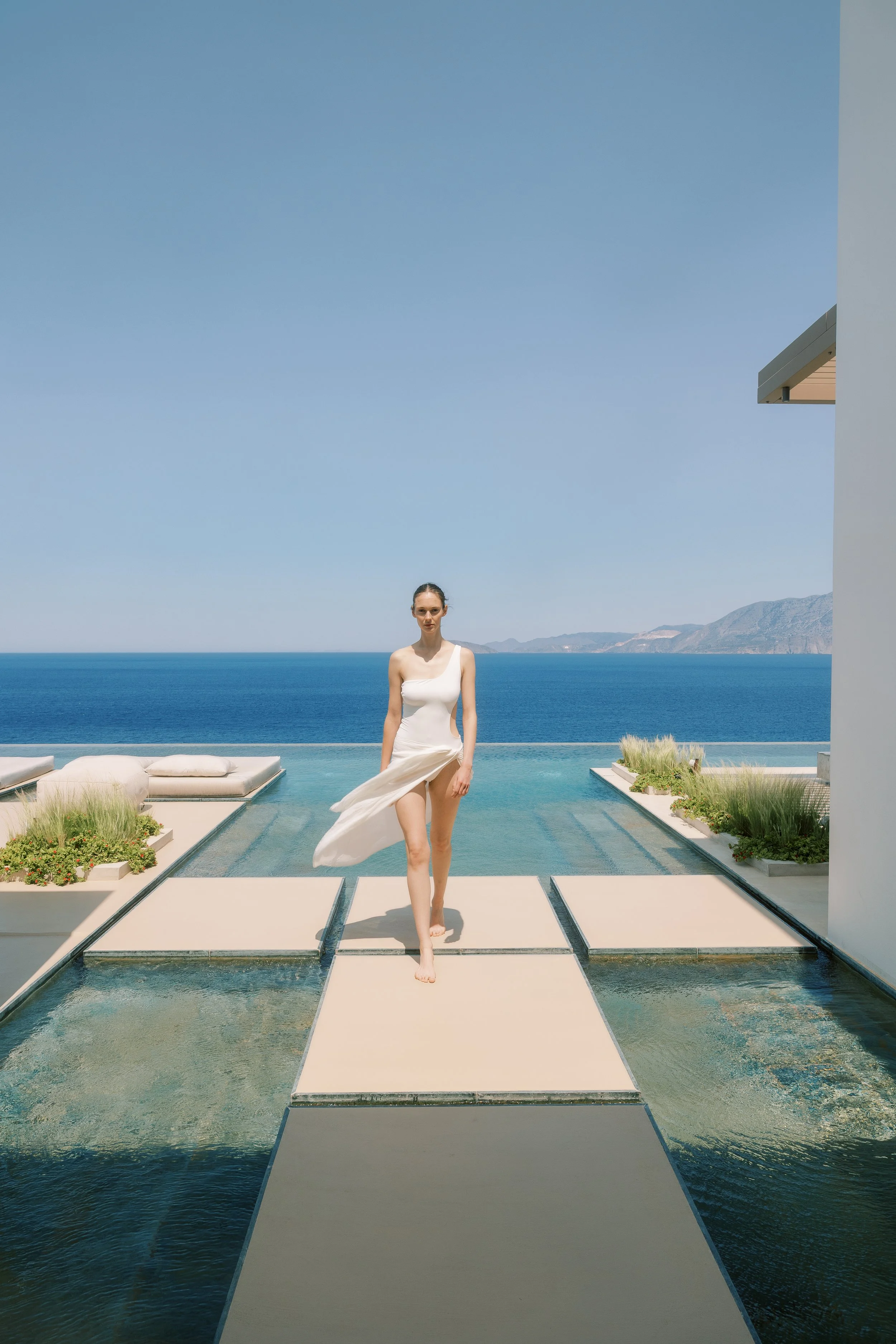 A woman walking on a narrow pathway over water with a modern building and mountains in the background, under a clear blue sky.