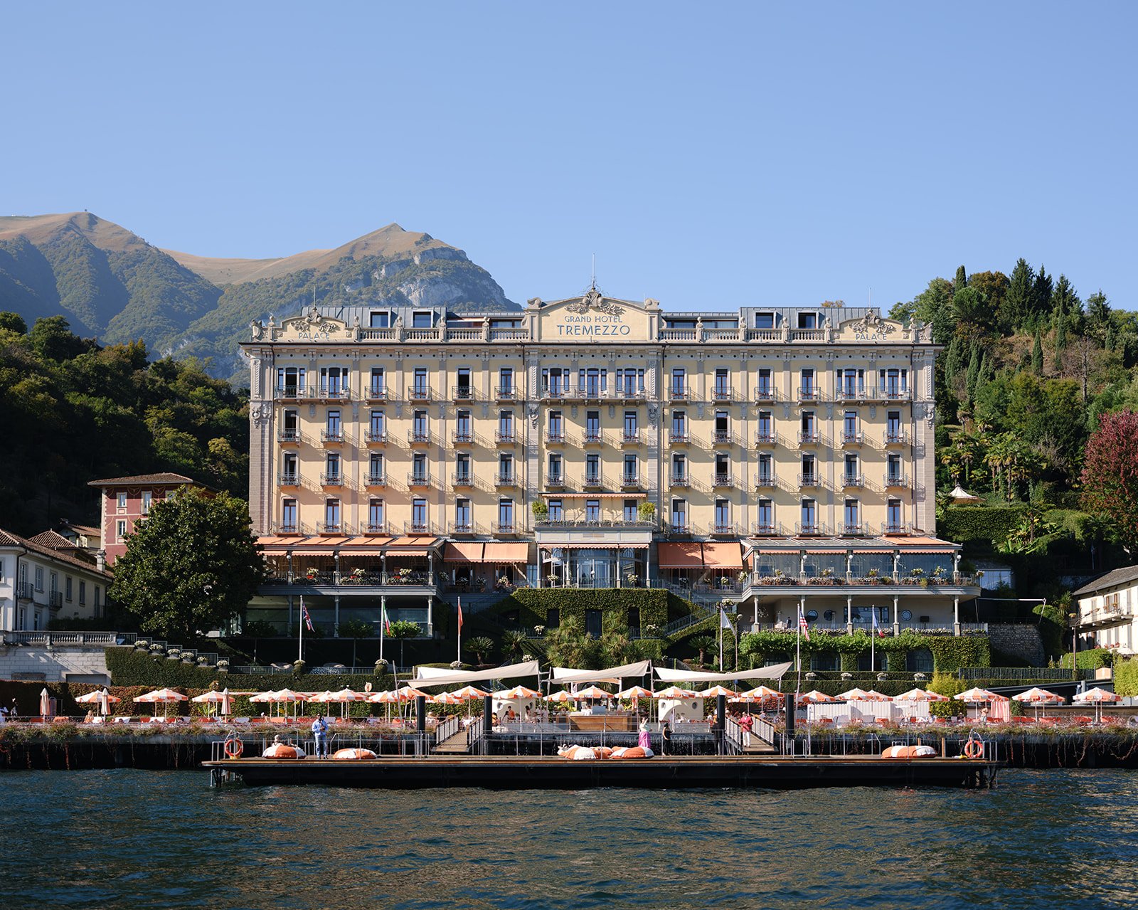 The Grand Hotel Tremezzo, a luxurious lakeside hotel with a classic architecture, set against a backdrop of mountains and greenery, with outdoor seating and umbrellas along the shoreline.