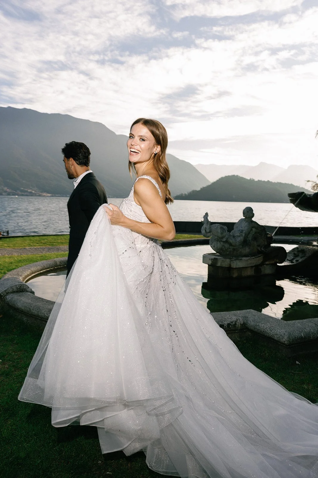 Bride smiling beside a historic fountain with Lake Como and the surrounding mountains behind her.