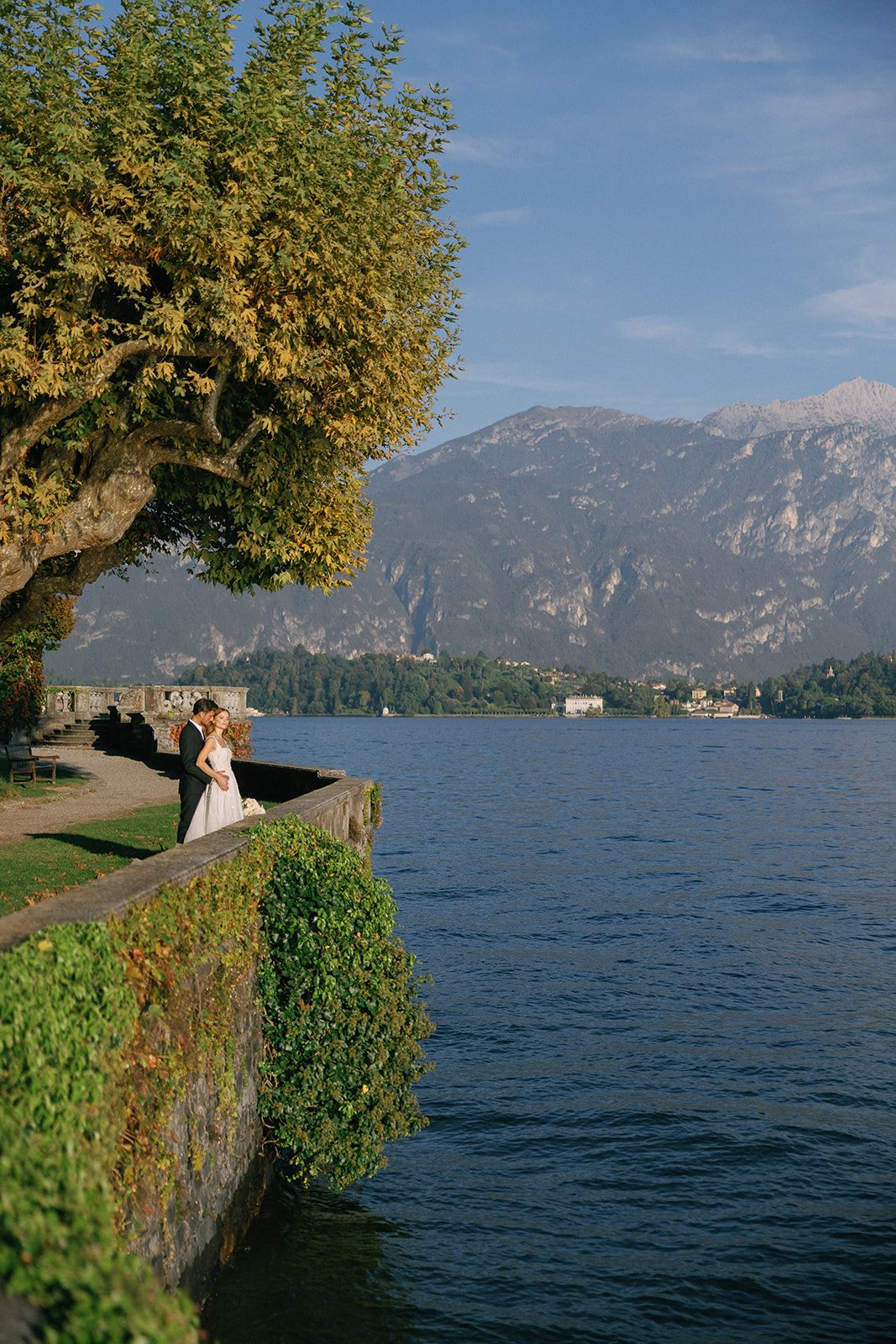 Couple embracing beneath a mature tree in a historic lakeside garden overlooking Lake Como.