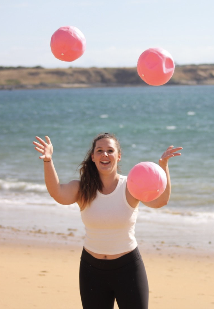 A woman playing with pink balls on a beach, with water and a distant shoreline in the background.