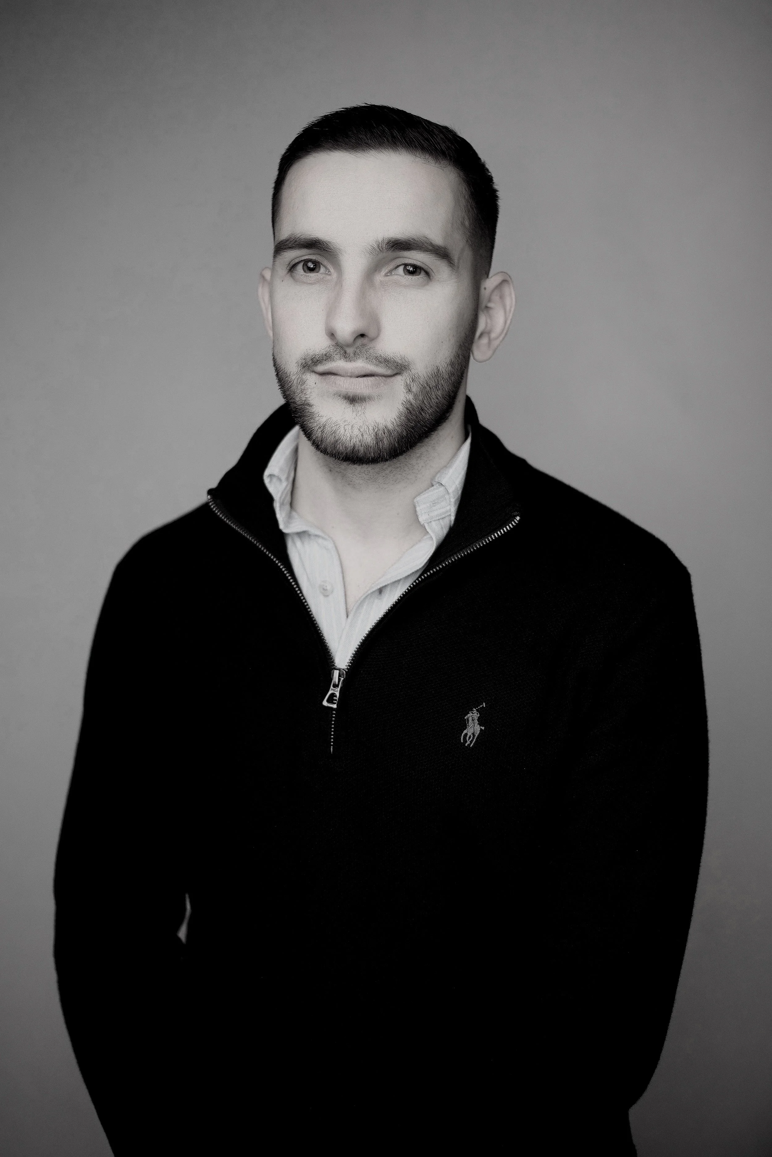 Black and white portrait of a young man with short dark hair, light skin, a beard, wearing a white collared shirt and a black zip-up jacket with a small Polo logo.
