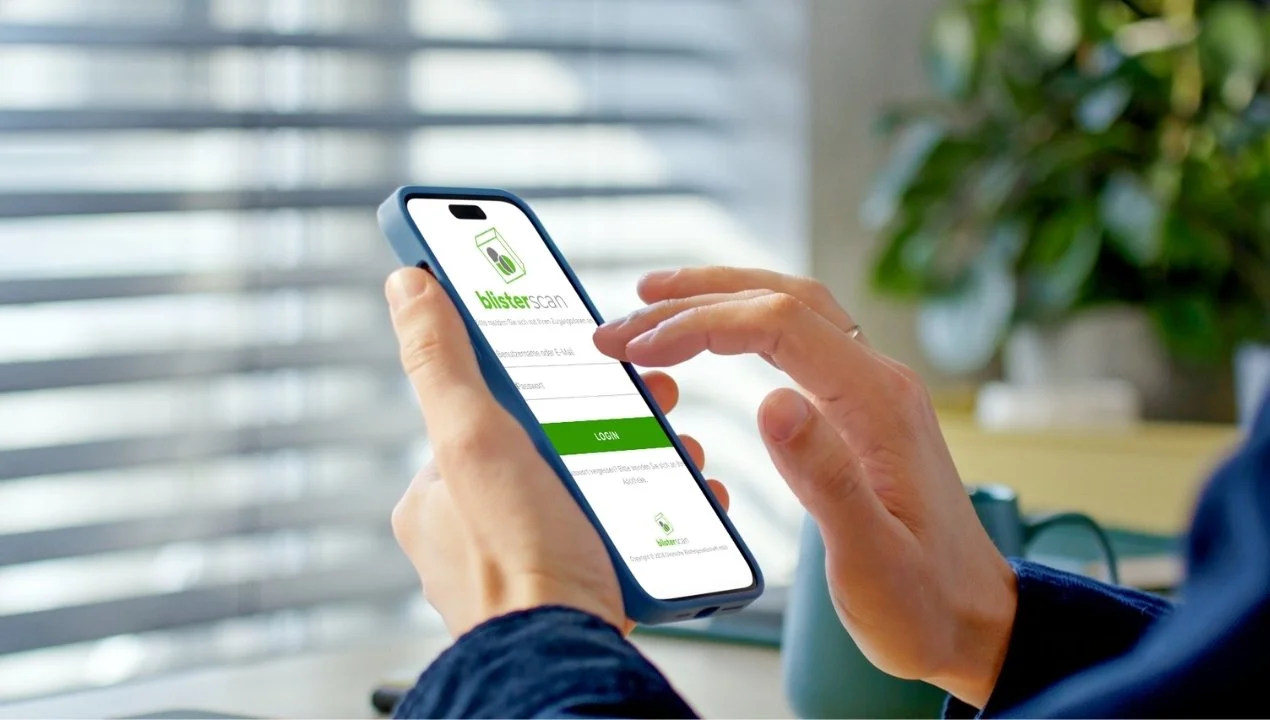 Eine Person hält ein Smartphone mit einer App namens 'blisters scan', die eine Login-Seite zeigt, vor einem Fenster mit horizontalen Jalousien und einer Topfpflanze im Hintergrund.