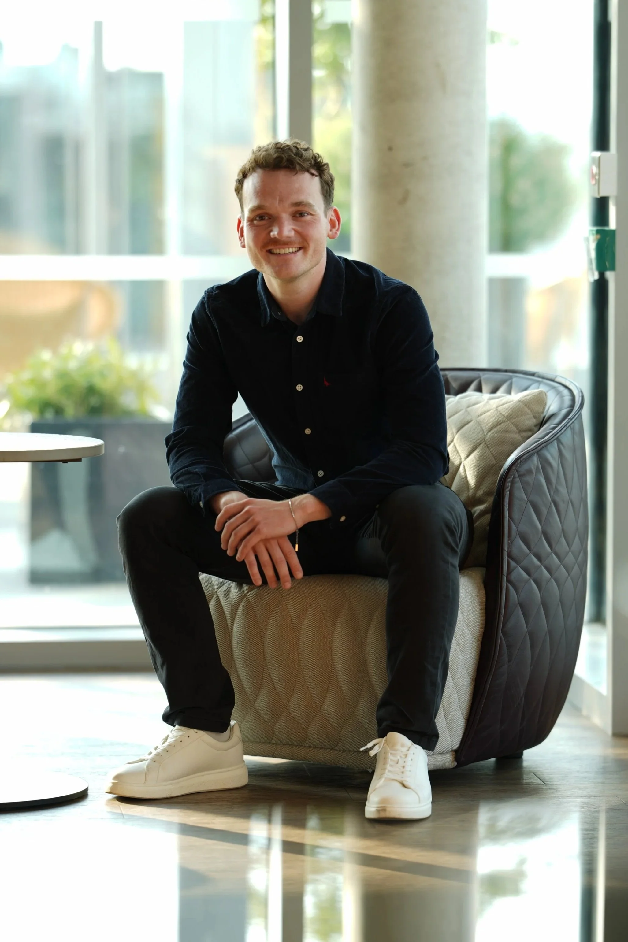A young man with curly brown hair and a smile sits in a modern lounge area near large windows, wearing a black shirt, black pants, and white sneakers.