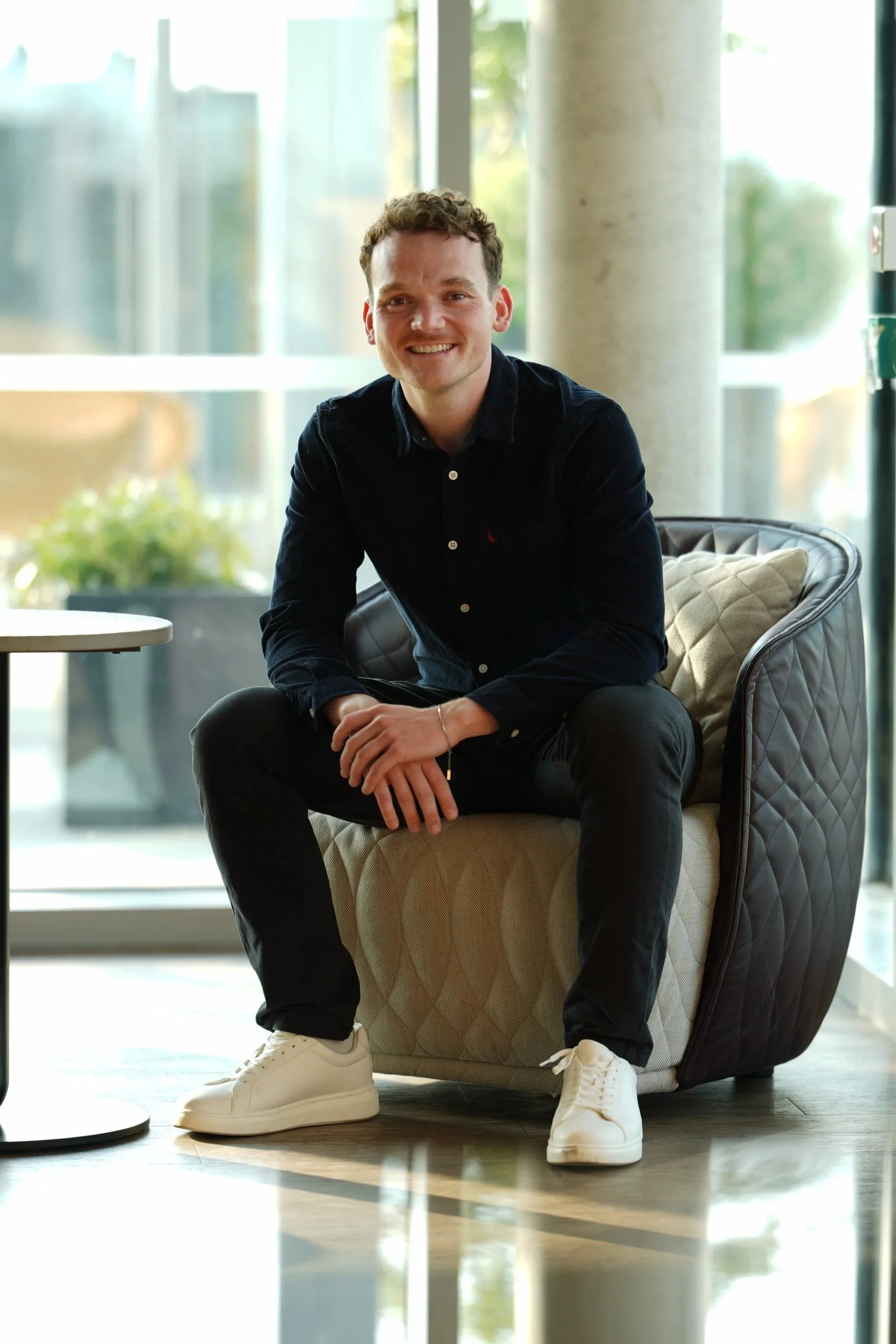 A young man sitting on a beige quilted chair, smiling, in a modern indoor setting with large windows, greenery outside, a small round table to his left, and a light-colored pillar behind him. He is wearing a black button-up shirt, black pants, and white sneakers.