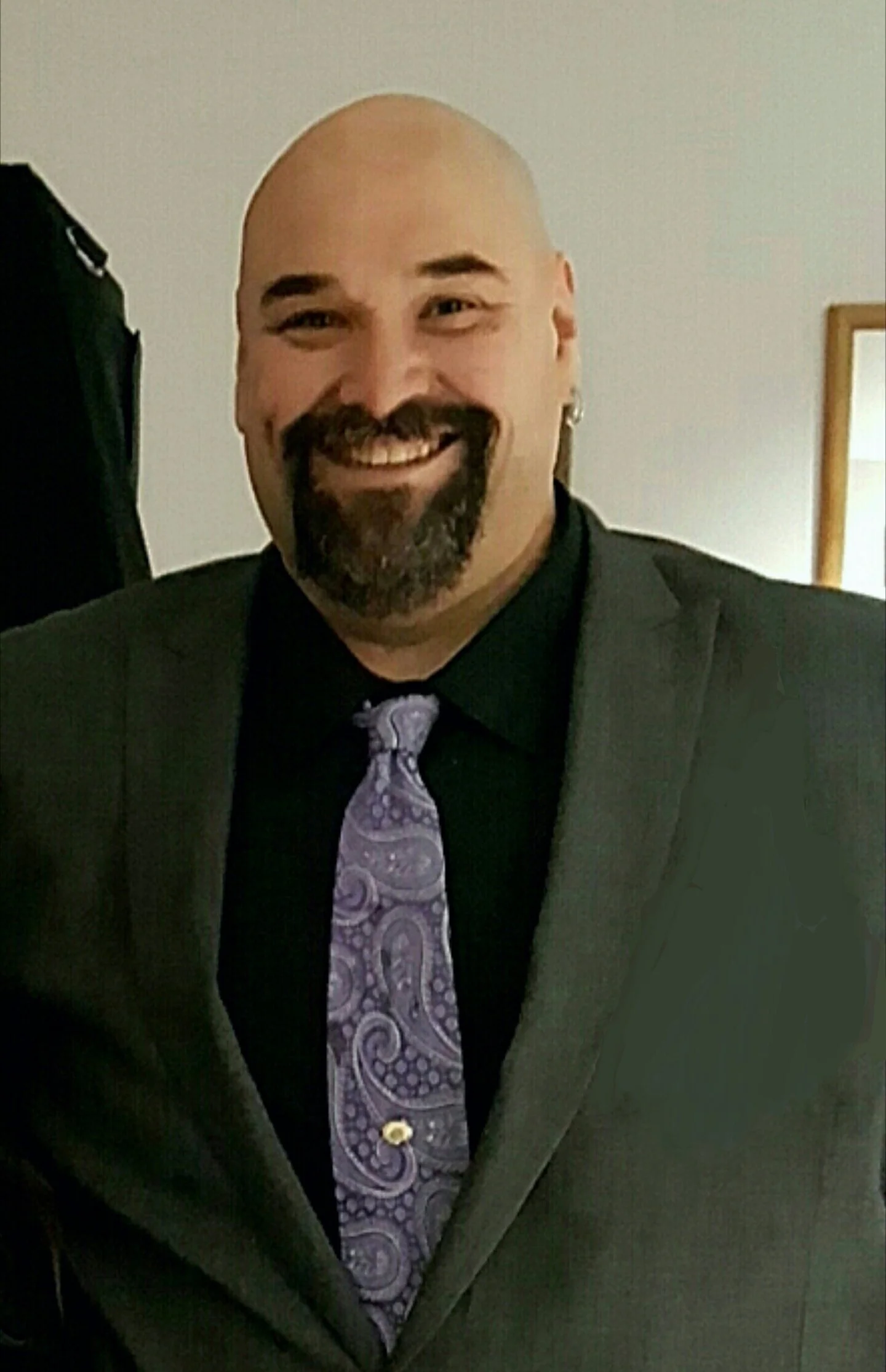 A bearded man with a shaved head smiling and wearing a dark suit, a lavender tie with a paisley pattern, and a yellow pin on his lapel.