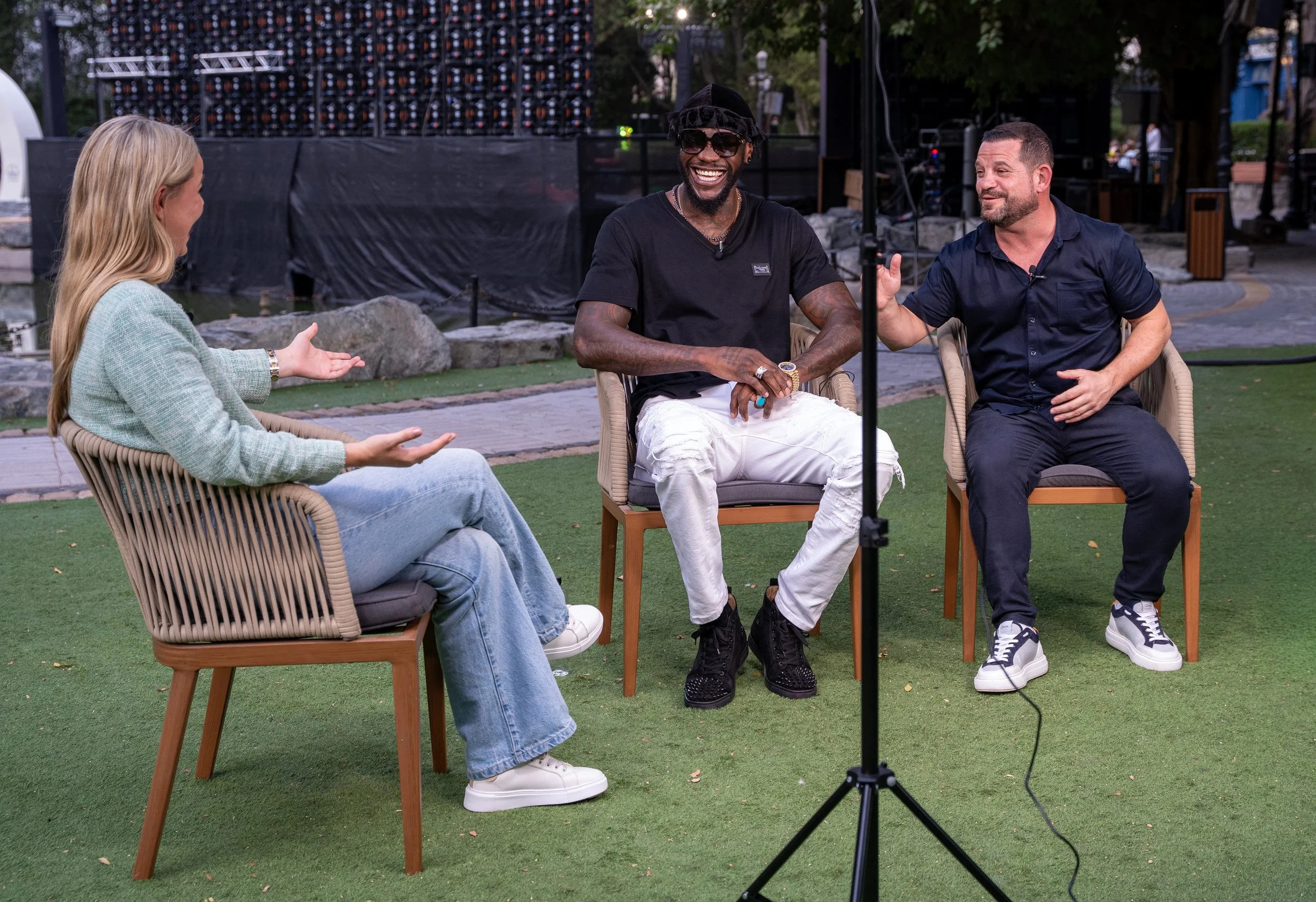 Three people sitting outdoors in a park, participating in an interview or discussion. One man is smiling broadly, wearing sunglasses and a black t-shirt. The woman on the left is gesturing with her hands, and the man on the right is listening and ges