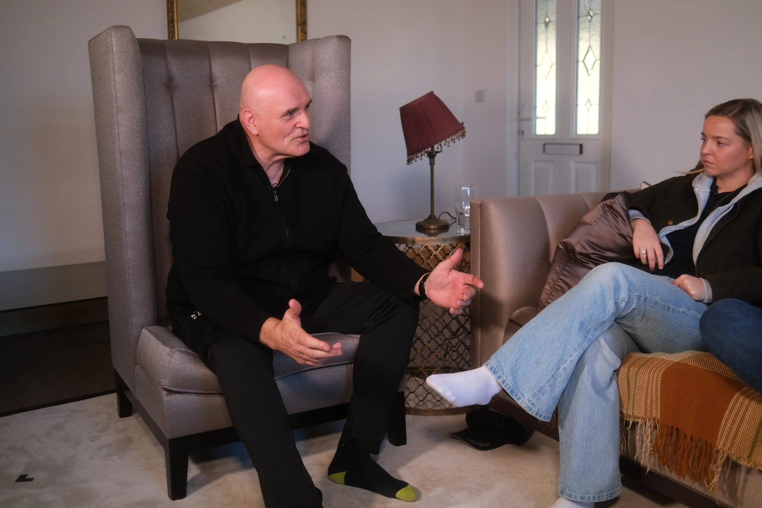 A bald man in a black jacket talking to a young woman sitting on a couch in a living room.