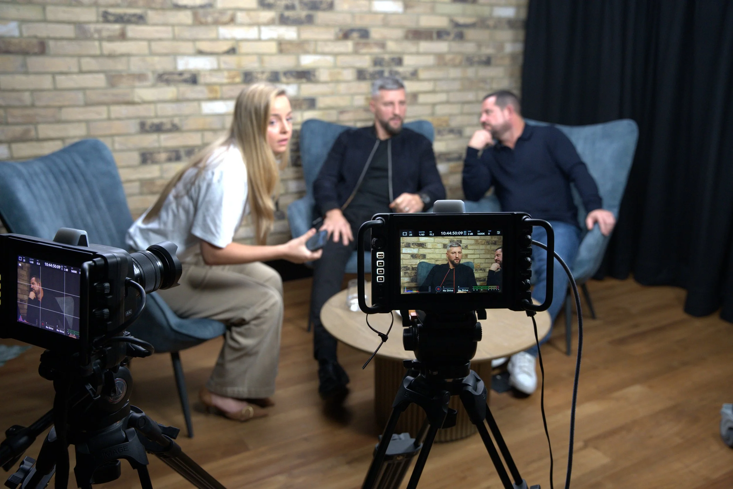 A woman and two men seated on a sofa, being filmed with professional cameras during an interview or talk show.