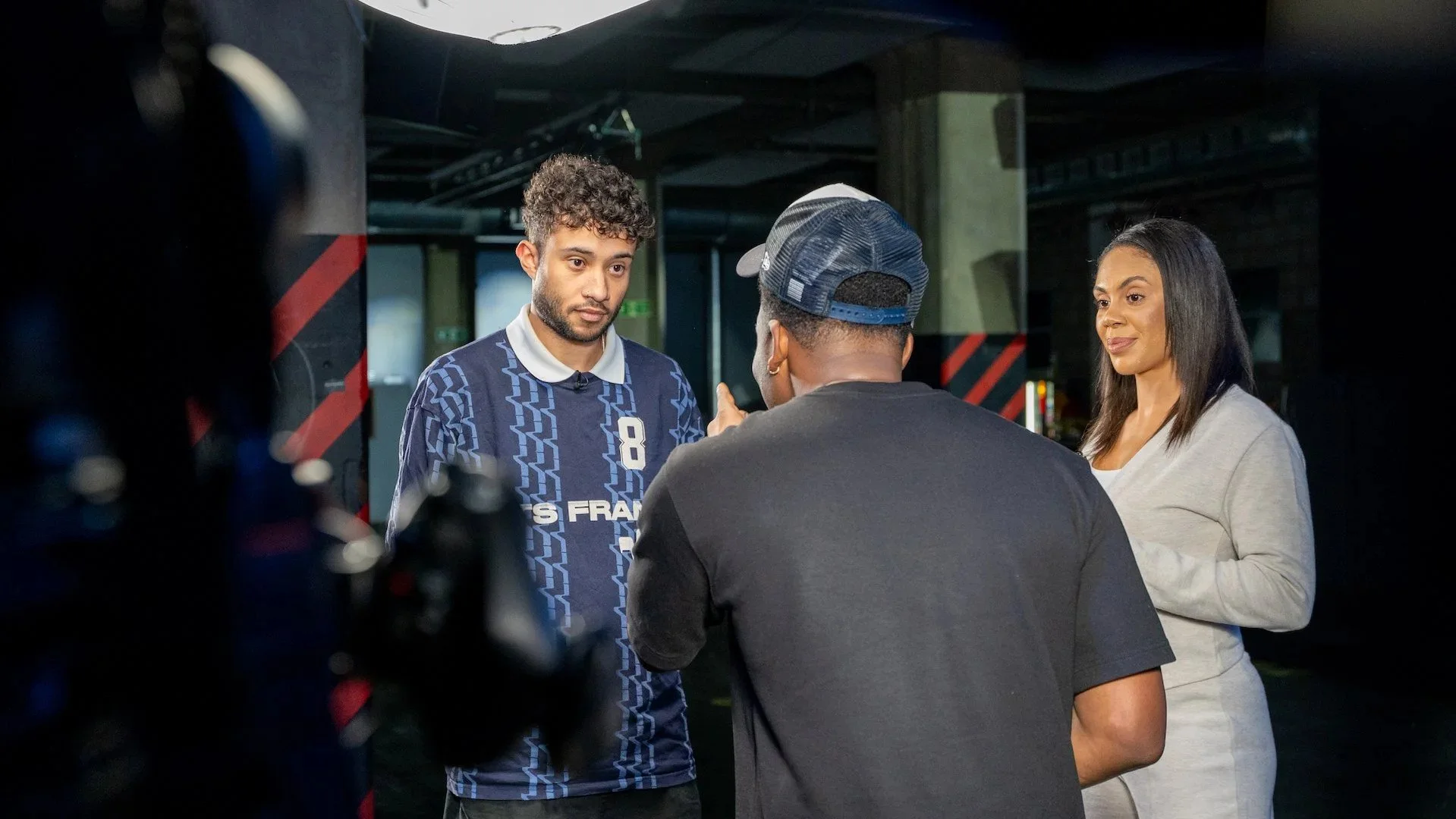 Three people having a discussion in an indoor setting, with a camera partially visible in the foreground.