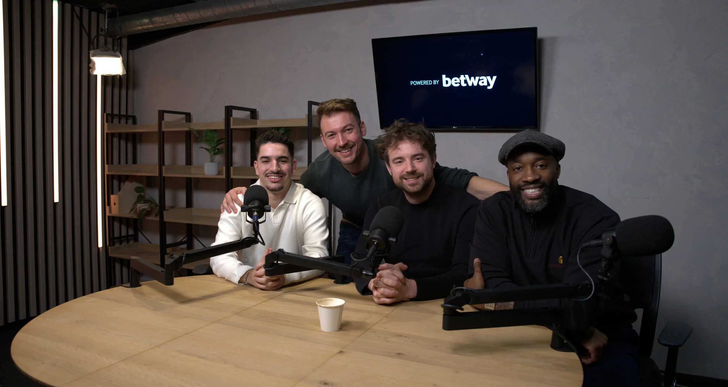 Four men sitting around a round wooden table with microphones in front of them, two are smiling and the other two are posing for the camera in a casual setting, with a large screen behind displaying 'Powered by Betway'.