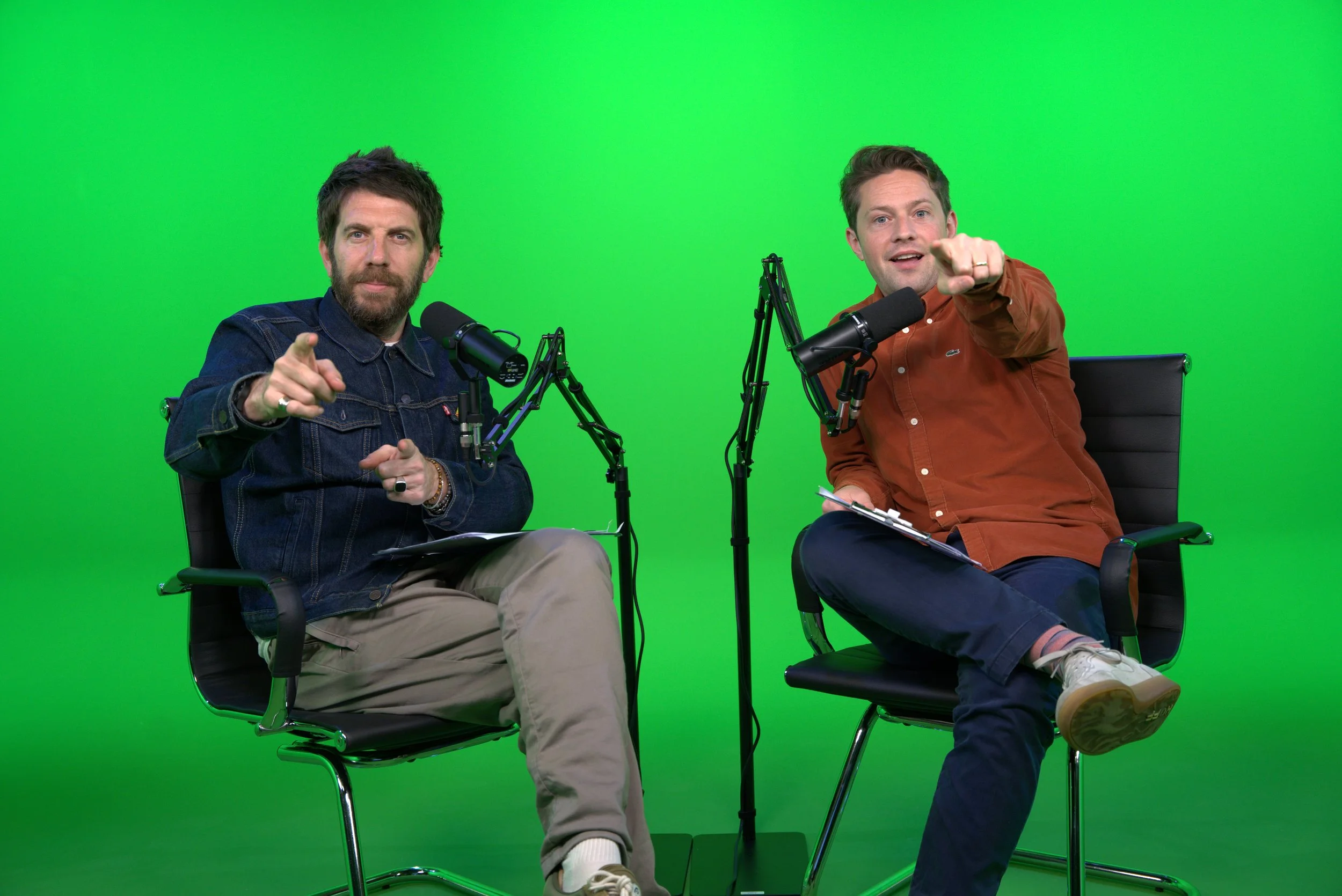 Two men sitting on black chairs with microphones in front of a green screen, pointing at the camera, one with a beard and denim jacket, the other in a brown shirt, holding papers.