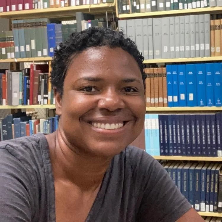 A smiling person with short curly black hair, wearing a gray T-shirt, sitting in front of bookshelves filled with various colored books in a library.