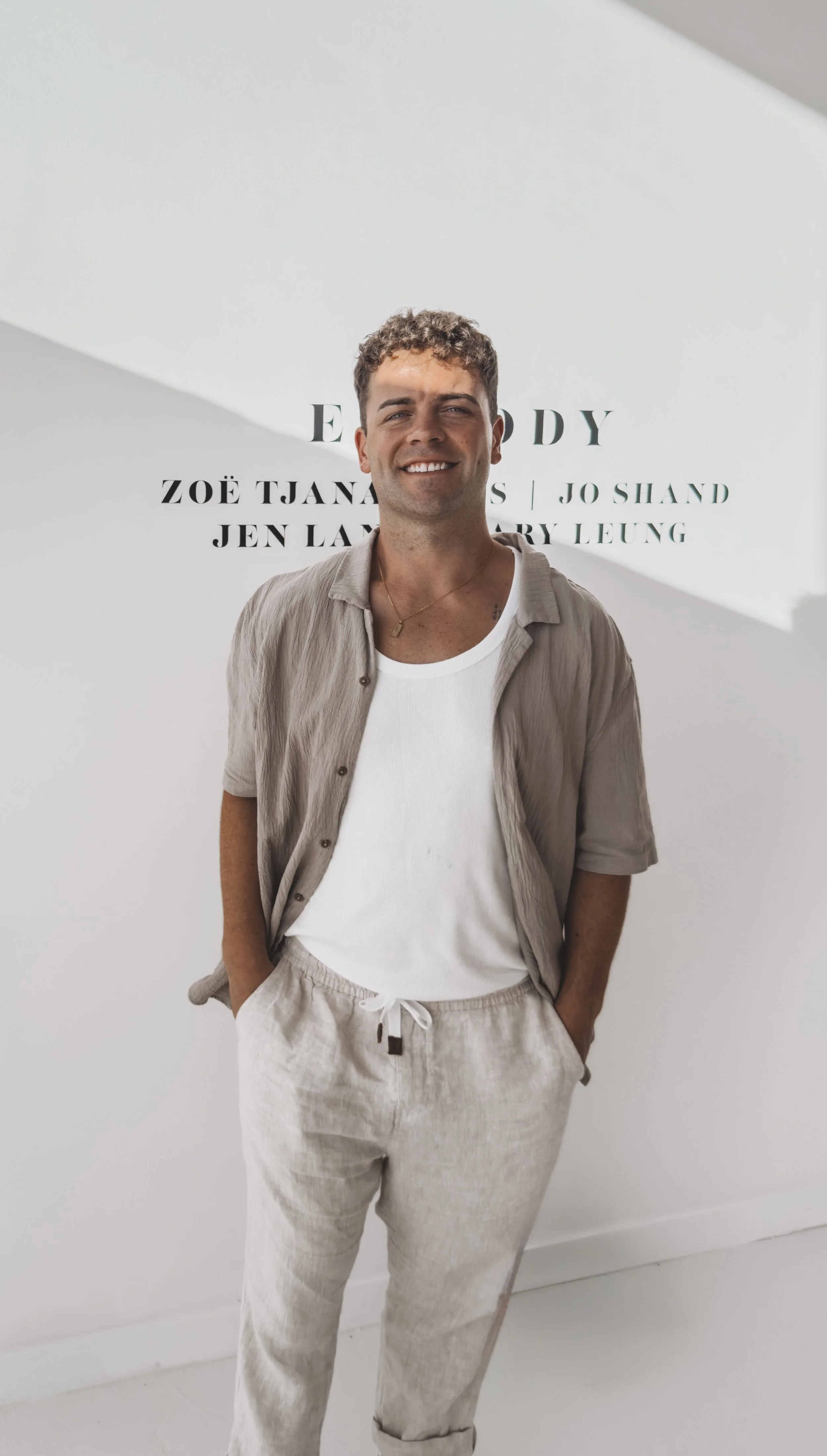 A young man with curly brown hair and a smile, wearing a beige shirt over a white t-shirt, beige drawstring pants, standing with hands in pockets in front of a white wall with black text.