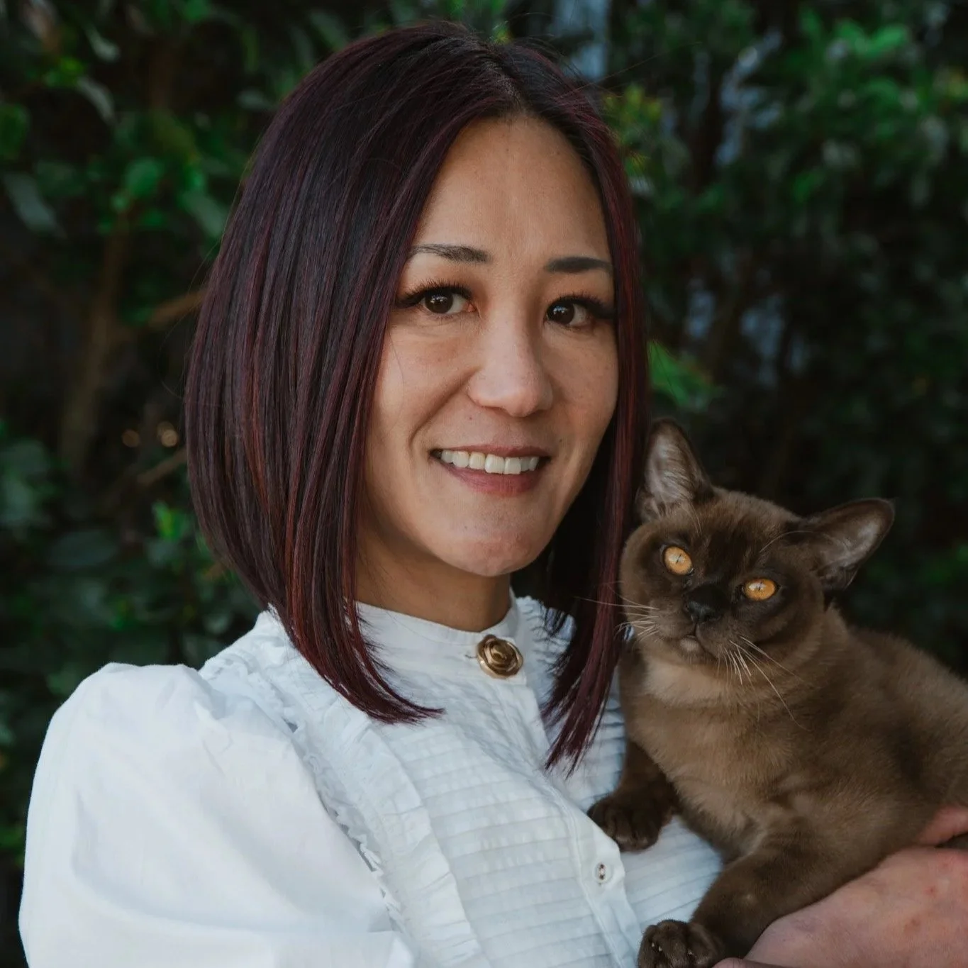 A woman with shoulder-length dark hair with red highlights, wearing a white top, holding a brown cat with yellow eyes, both smiling outdoors with green foliage in the background.
