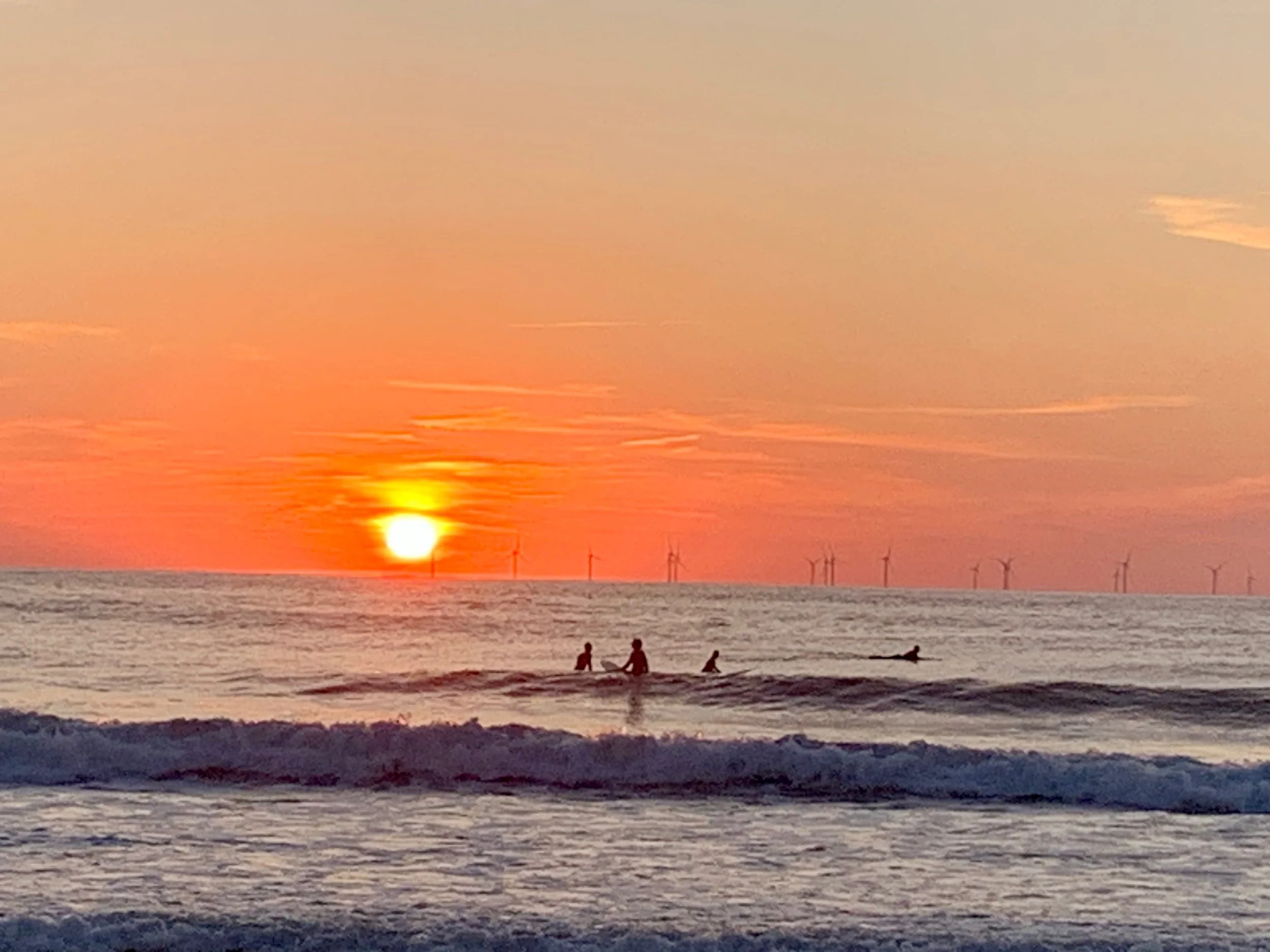 Three surfers sitting on their boards in front of an orange sunset over the North Sea, wind turbines along the horizon
