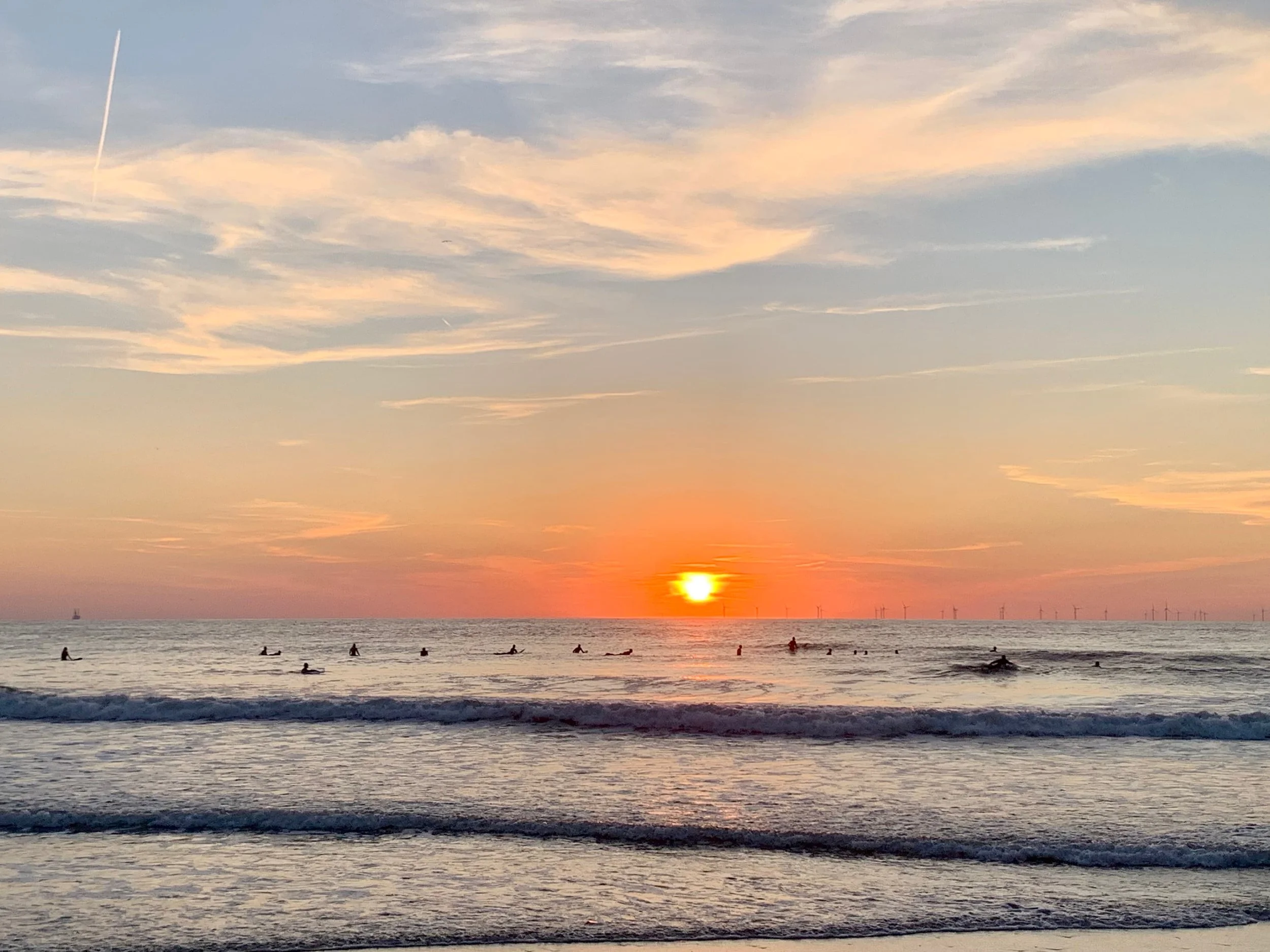 Alt: Surfers floating in the North Sea at sunset near The Hague, with offshore wind turbines on the horizon