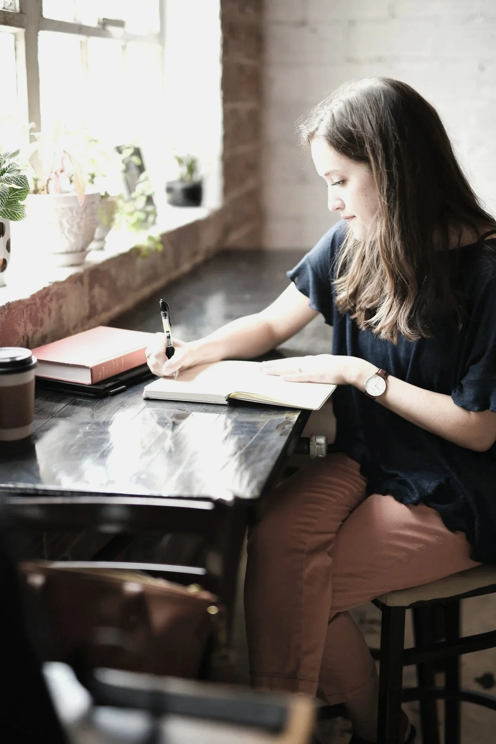 A woman with long brown hair, wearing a black shirt and blush pants, is sitting at a black table with a notebook and books in front of her, writing with a pen, near a window with sunlight, potted plants, and a brick wall in the background.