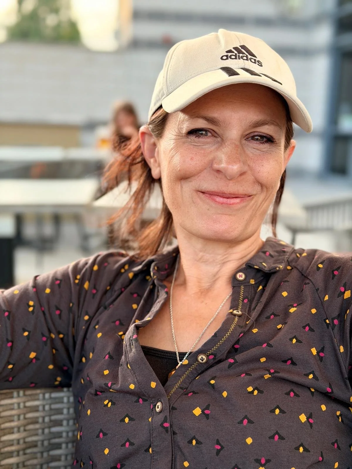 A woman smiling outdoors, wearing a beige Adidas cap and a dark shirt with small pink, yellow, and black geometric patterns.