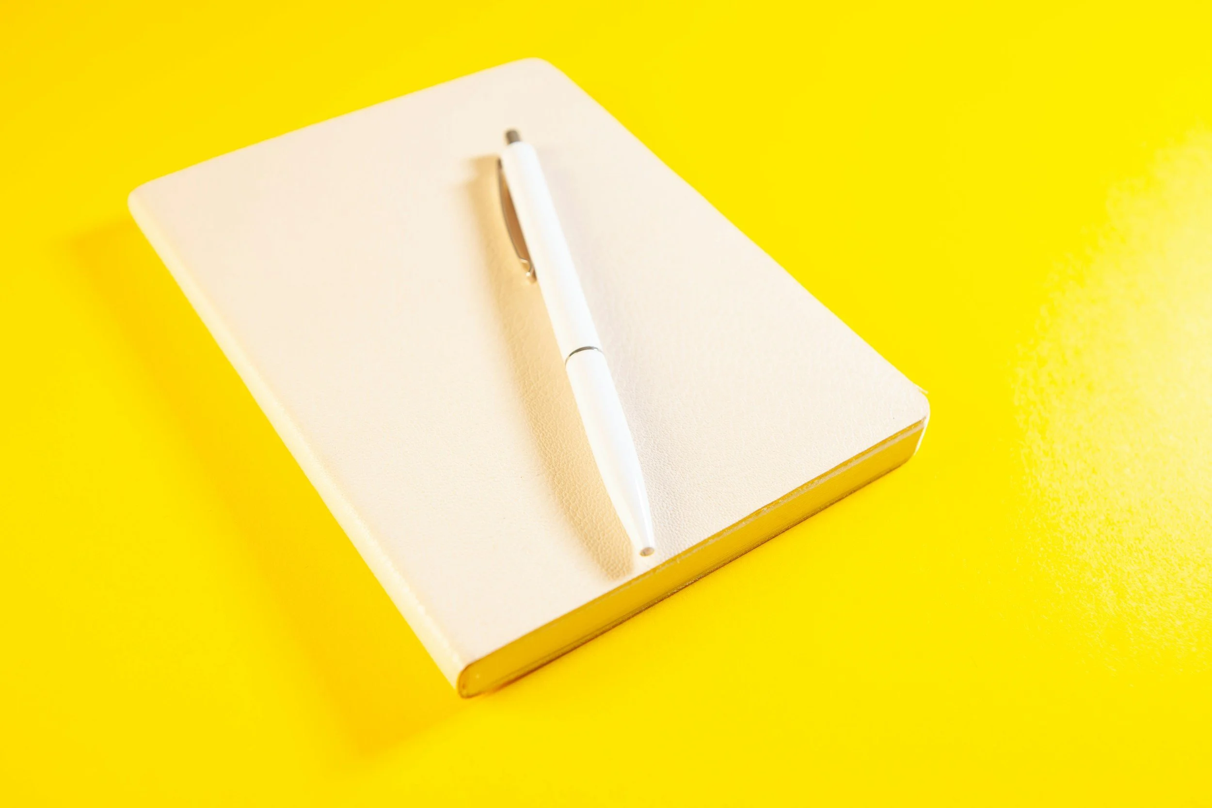 An open notebook with blank pages and a white pen resting on top, placed on a bright yellow surface.