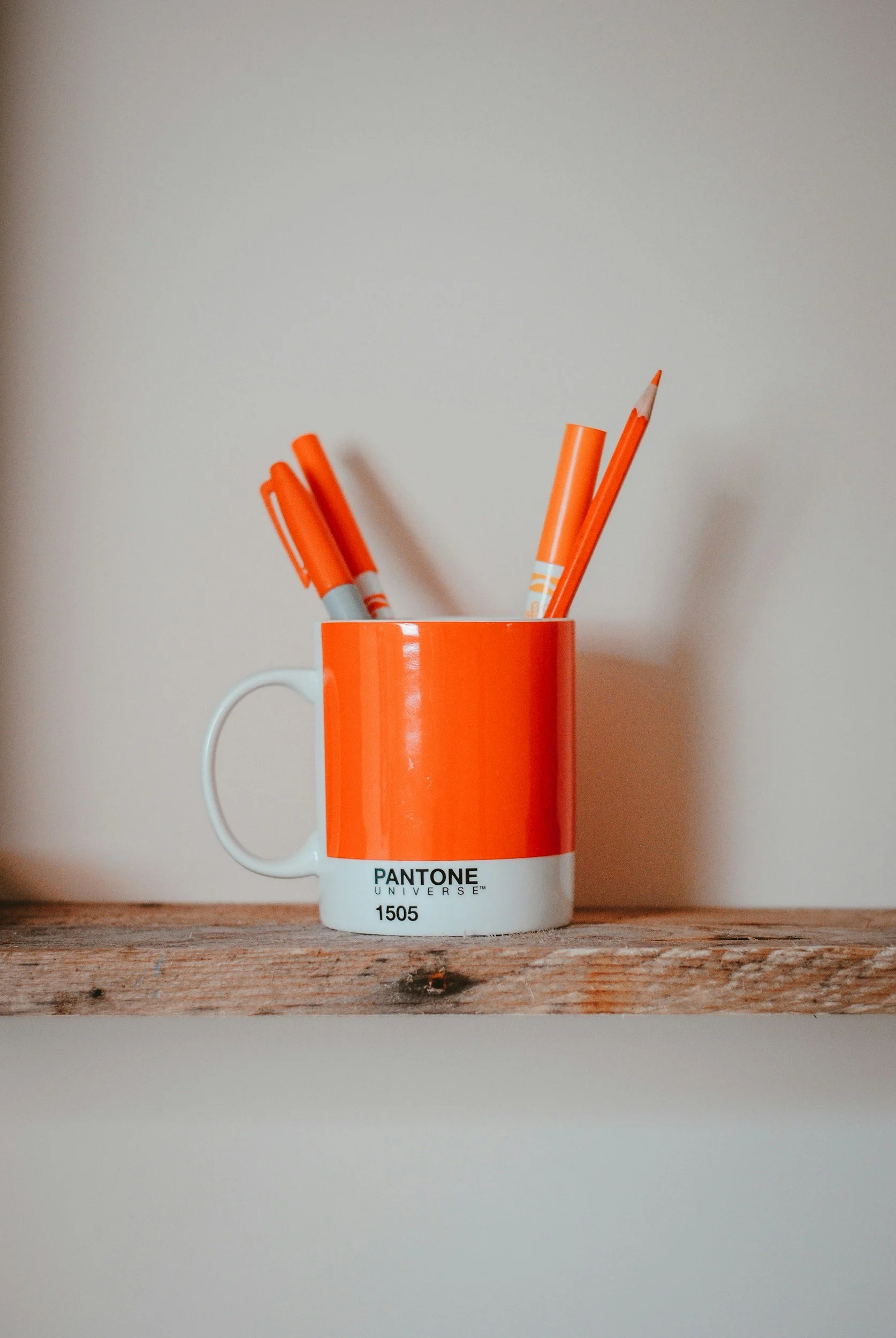 A white ceramic mug with an orange Pantone color design and the words "Pantone Universe 1505" printed on it, holding orange markers and a colored pencil, placed on a wooden surface against a plain background.