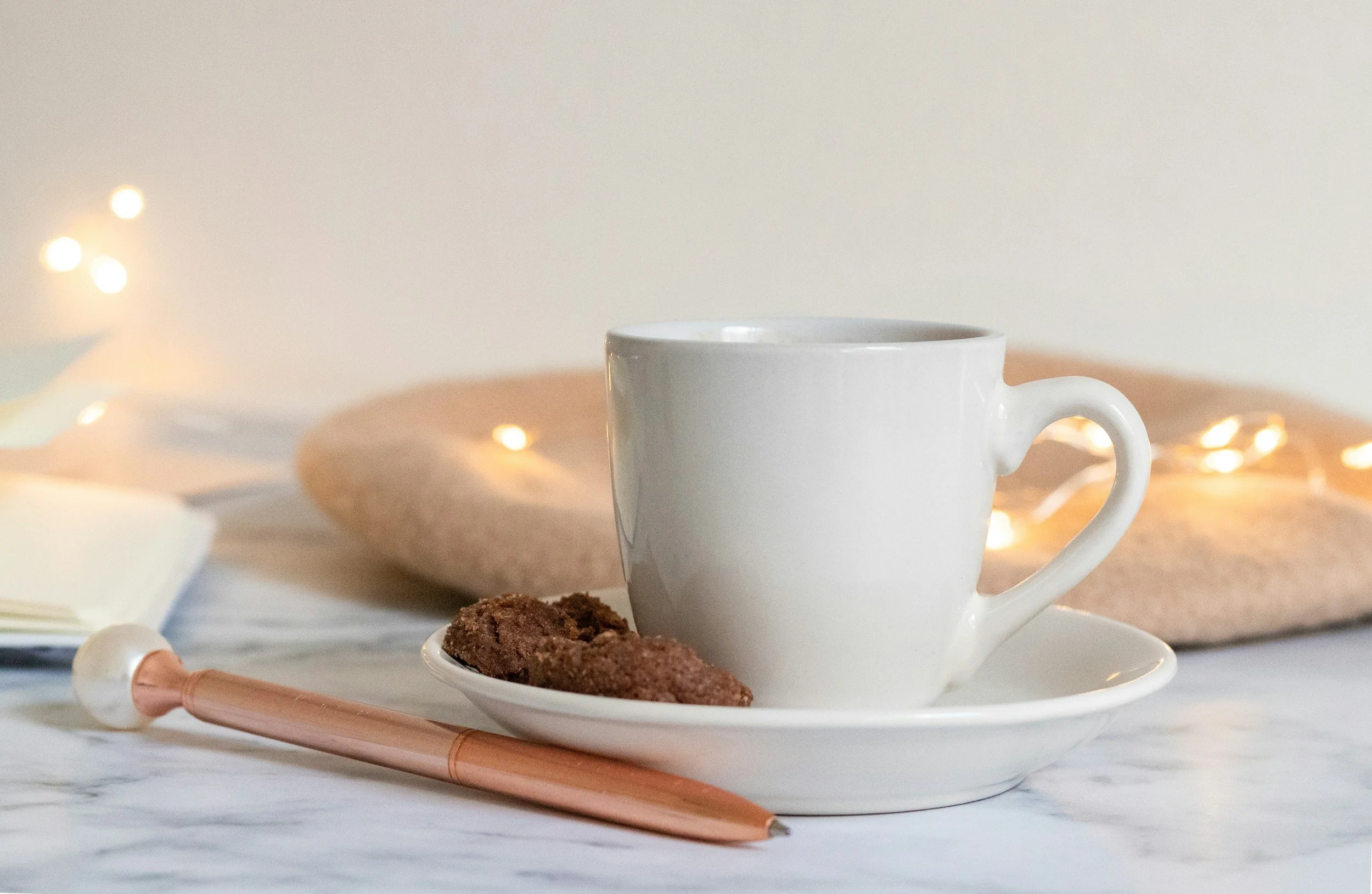A white coffee mug on a saucer with oatmeal cookies on a white plate, set on a table with a marble pattern, with blurred warm fairy lights in the background.