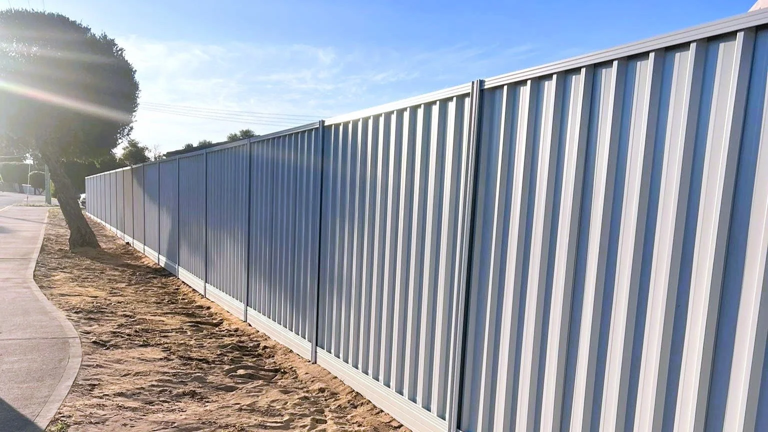 A metal fence runs alongside a sidewalk with a tree on the left, under a clear blue sky with the sun shining.
