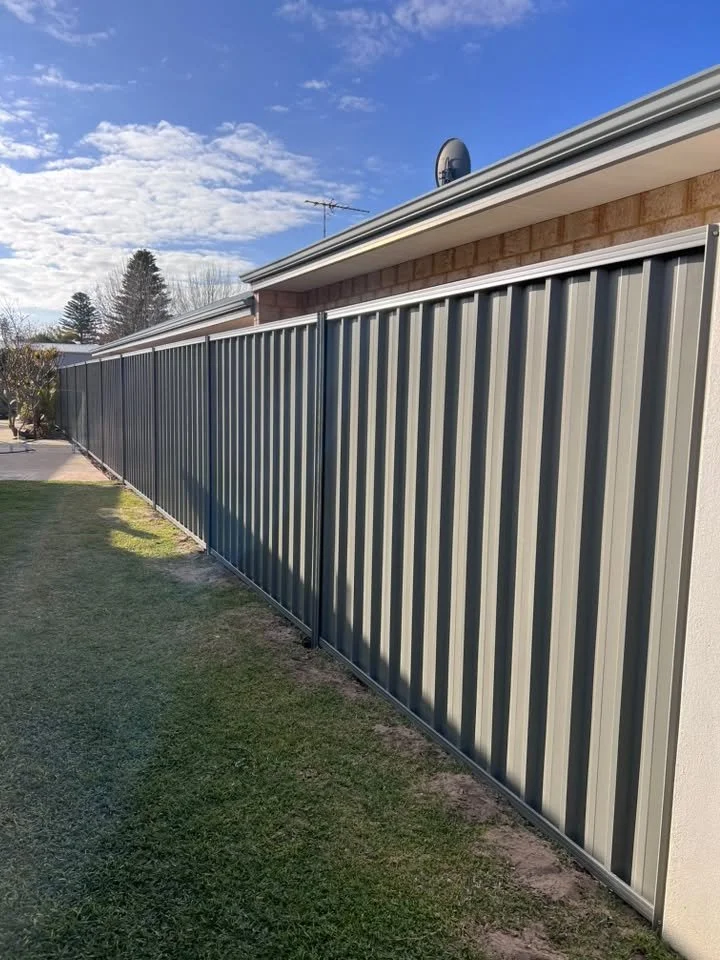 A long metal fence runs alongside a house with a brick exterior and a satellite dish. The yard has grass and the sky is partly cloudy.