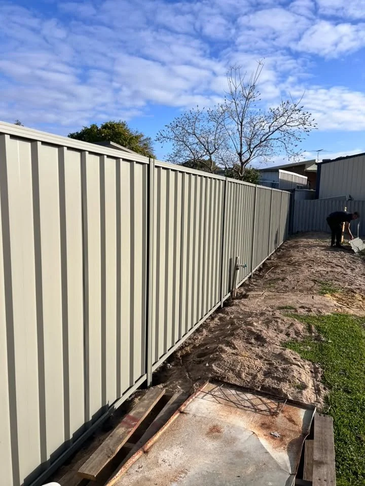 A beige metal fence running along a backyard, with a worker in the background watering or cleaning near a house. The sky is partly cloudy, and there are some leafless trees in the distance.