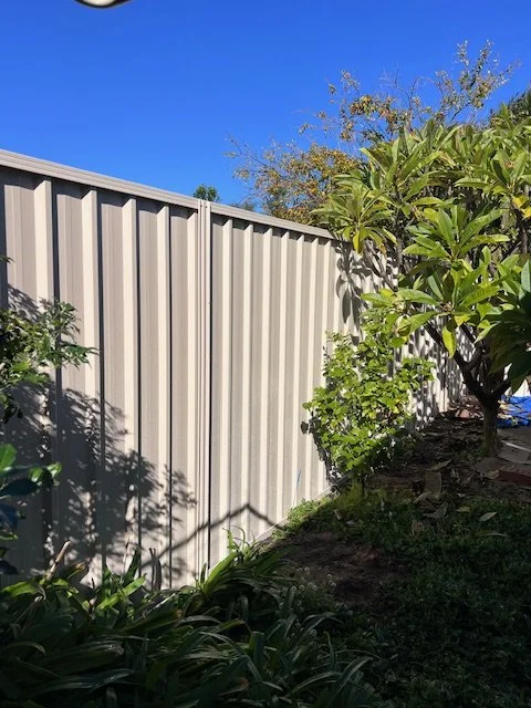 A beige metal privacy fence with shadows cast by nearby trees, adjacent to a lush garden with green plants and a tree, under a clear blue sky.