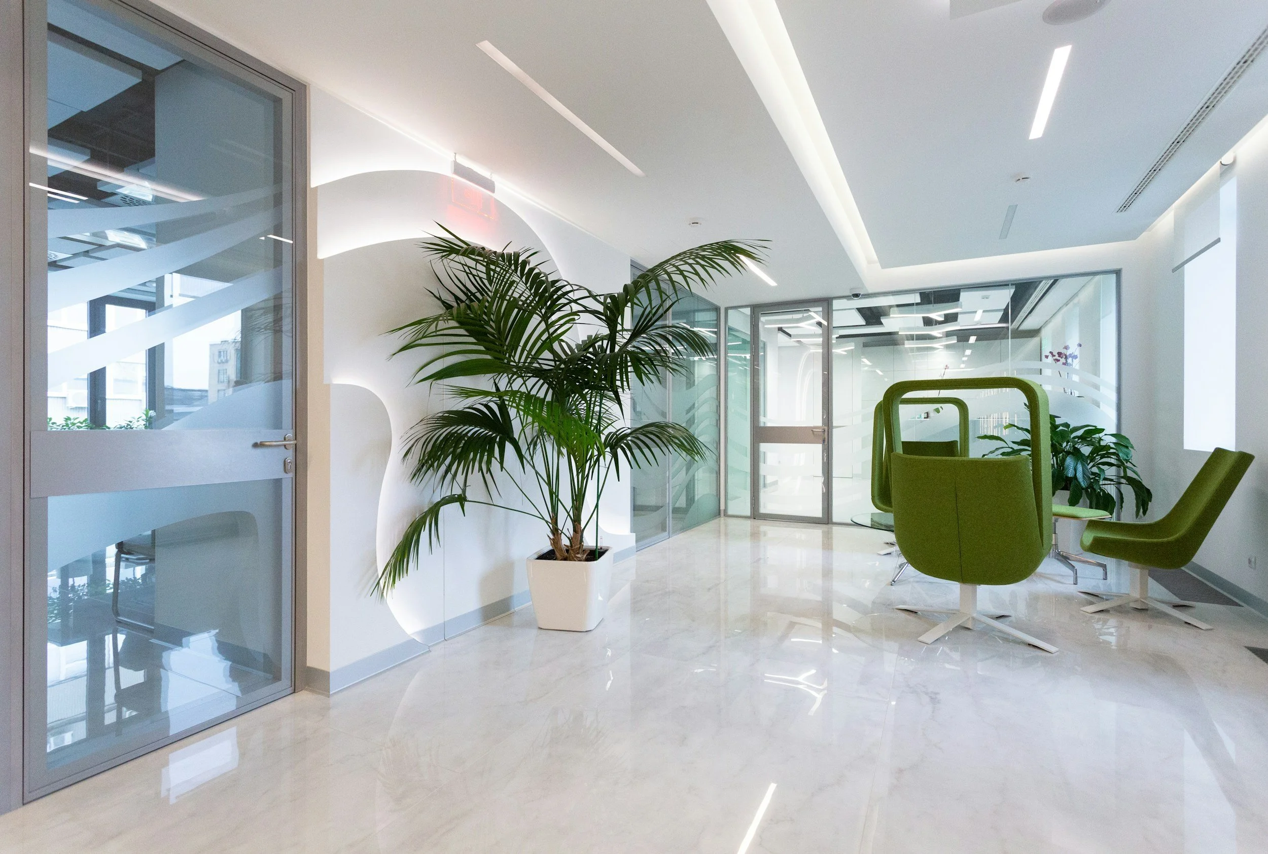 Modern office lobby with green chairs, potted plant, glass walls, and sleek lighting fixtures.