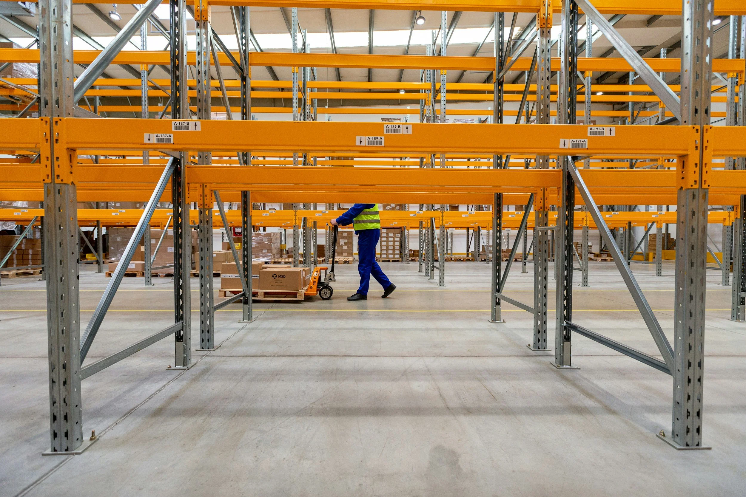 Warehouse storage area with metal shelving racks, some labeled with signs. A worker in a high-visibility vest is rolling a box on a cart in the aisle.