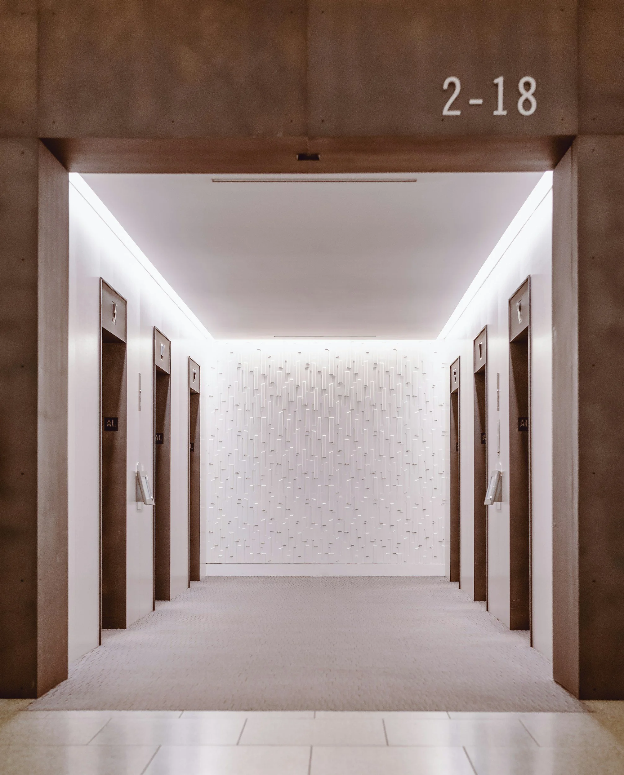 Inside an elevator lobby with a modern, clean design, featuring white walls with vertical textured patterns, and numbered signs 2-18 above elevator doors on both sides, with light-colored tiles on the floor and a textured white wall at the end.