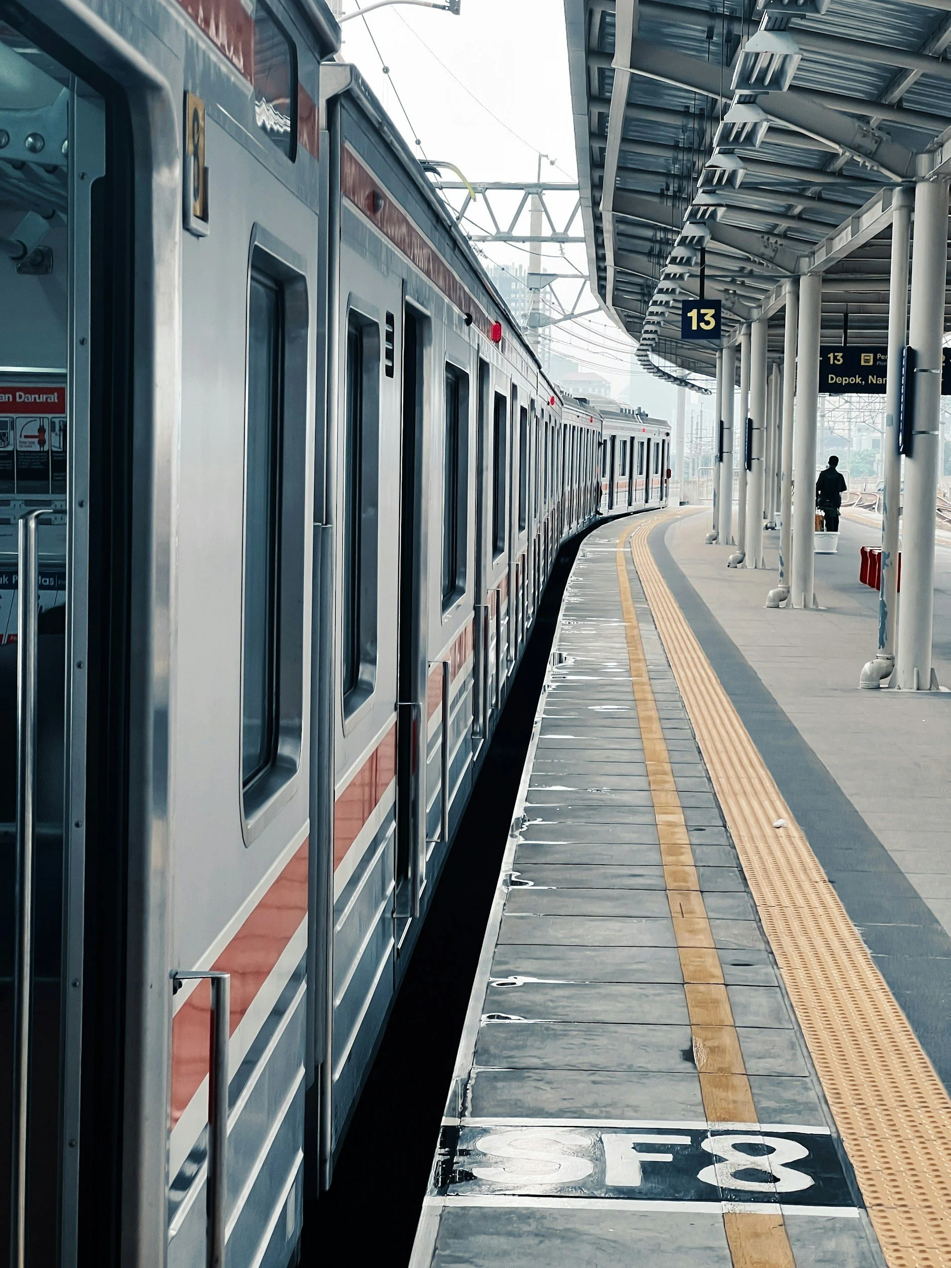 An electric train on a platform at a train station, with a person walking in the distance. There are overhead signs and shelter structures.