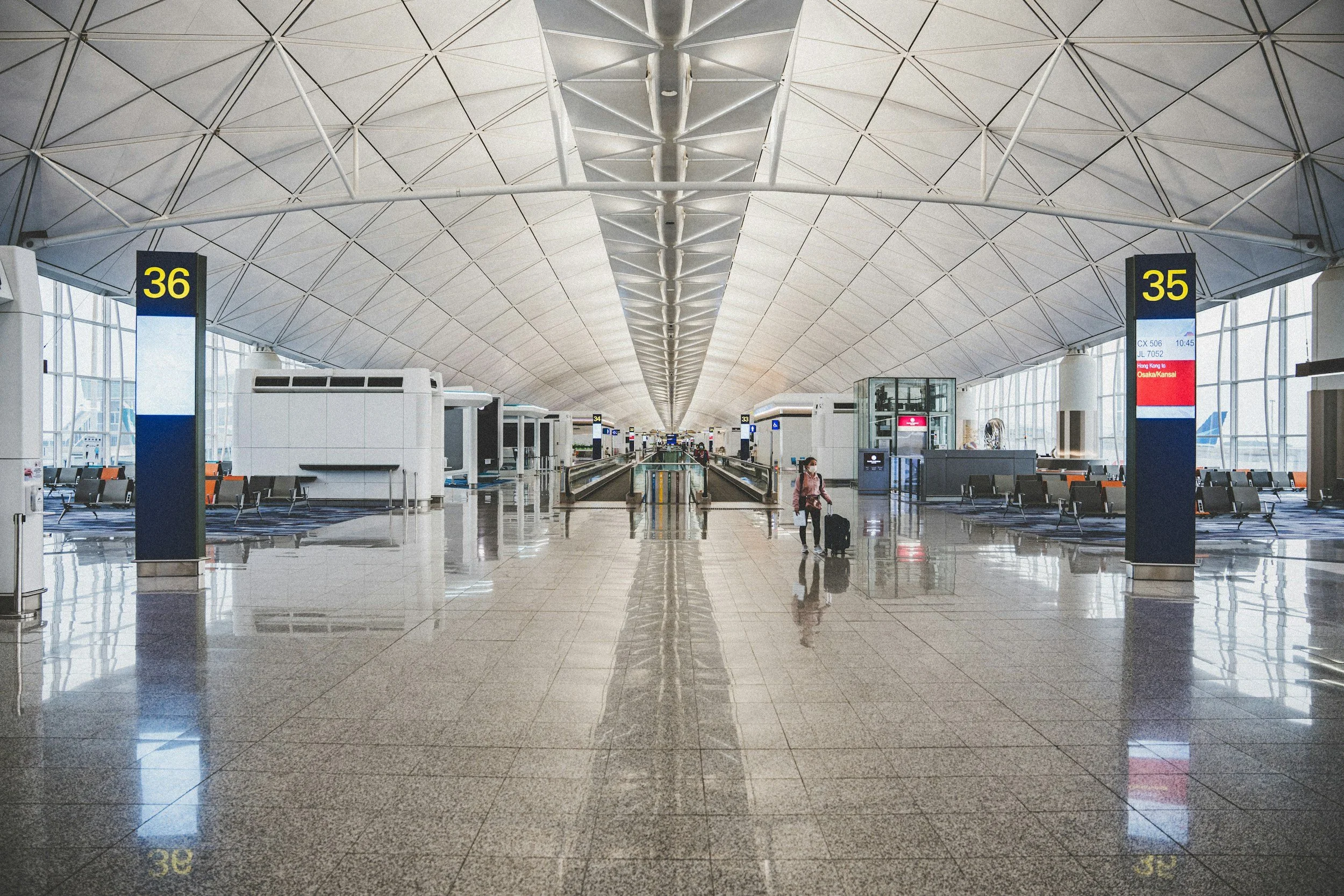 Interior of an airport terminal with high, curved ceiling and large windows, mostly empty with a few travelers and signs showing gate numbers 35 and 36.