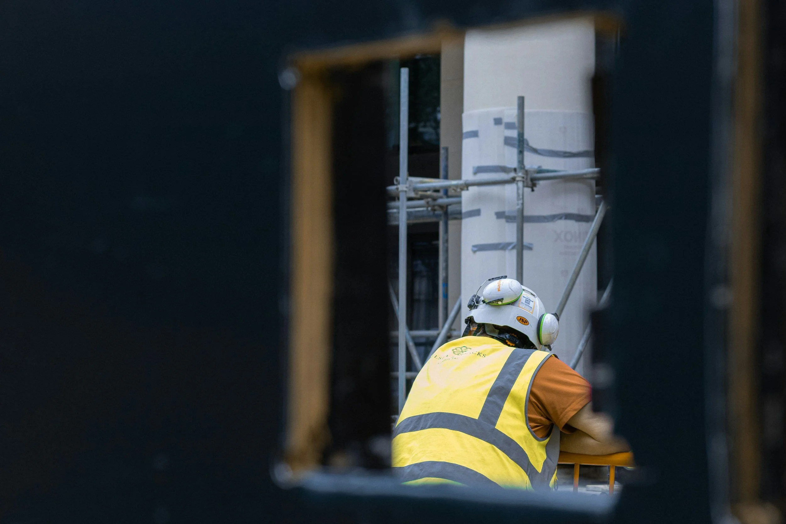 Construction worker wearing a yellow safety vest, white helmet, and orange shirt working on a building site, seen through a gap in the framing.