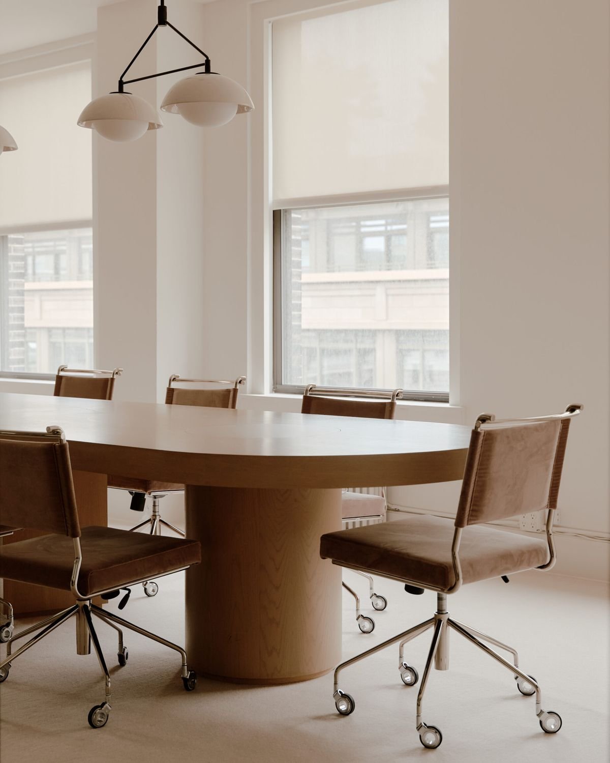 Modern conference room with a round wooden table, four brown leather chairs on caster wheels, and large windows with a city view, illuminated by natural light and a ceiling light fixture.