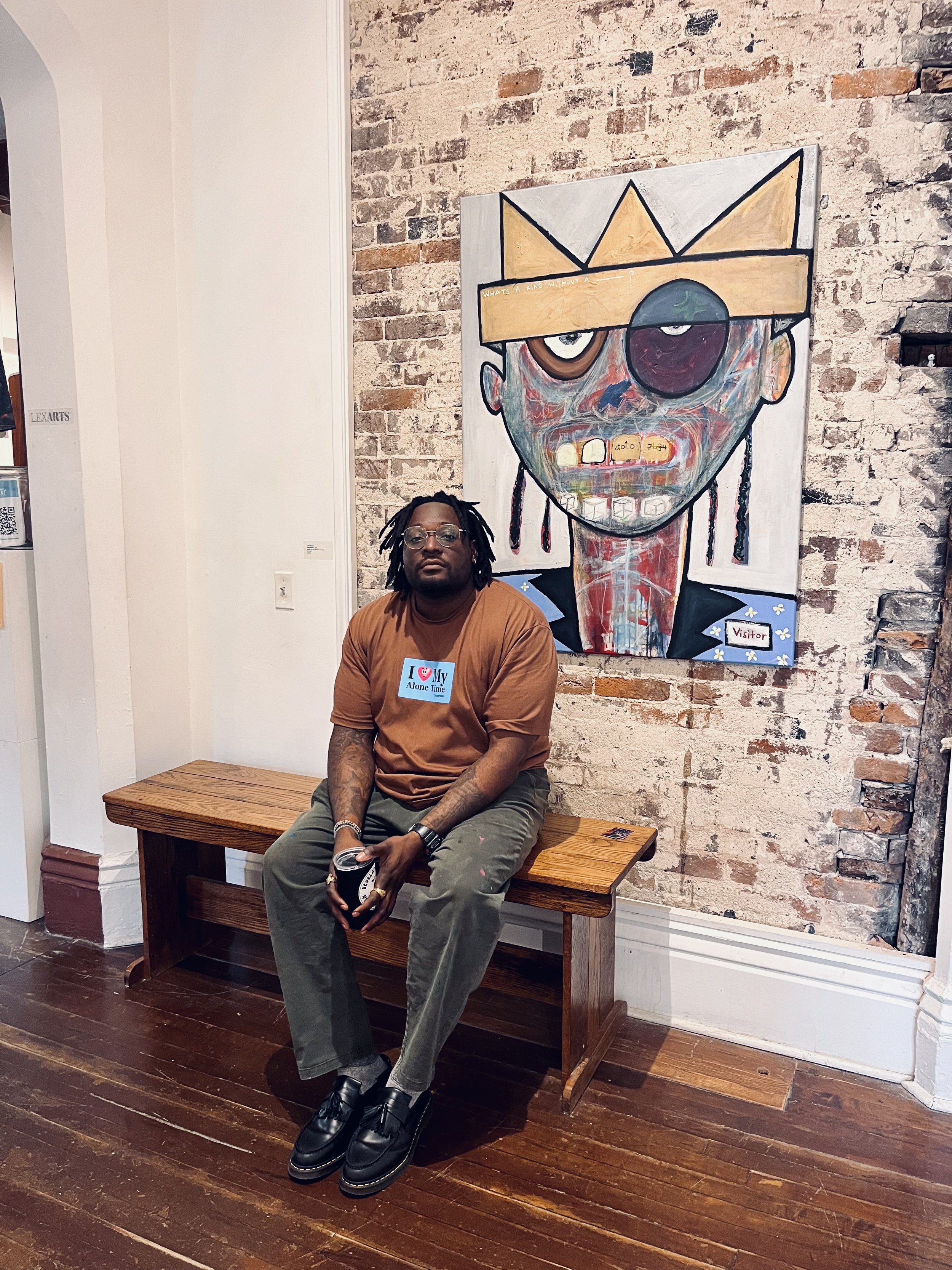 A man with glasses and dreadlocks sitting on a wooden bench in front of an abstract portrait of a face, painted on a brick wall. The man is wearing a brown shirt with a sticker that says 'I ❤️ My Alone Time' and green pants.