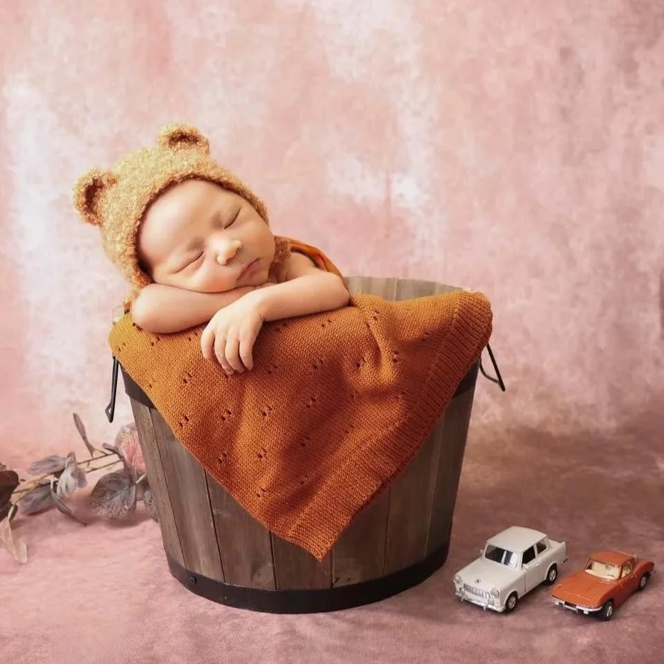 Sleeping baby with a bear hat, resting on an orange blanket inside a wooden bucket, with toy cars beside it against a pink background.