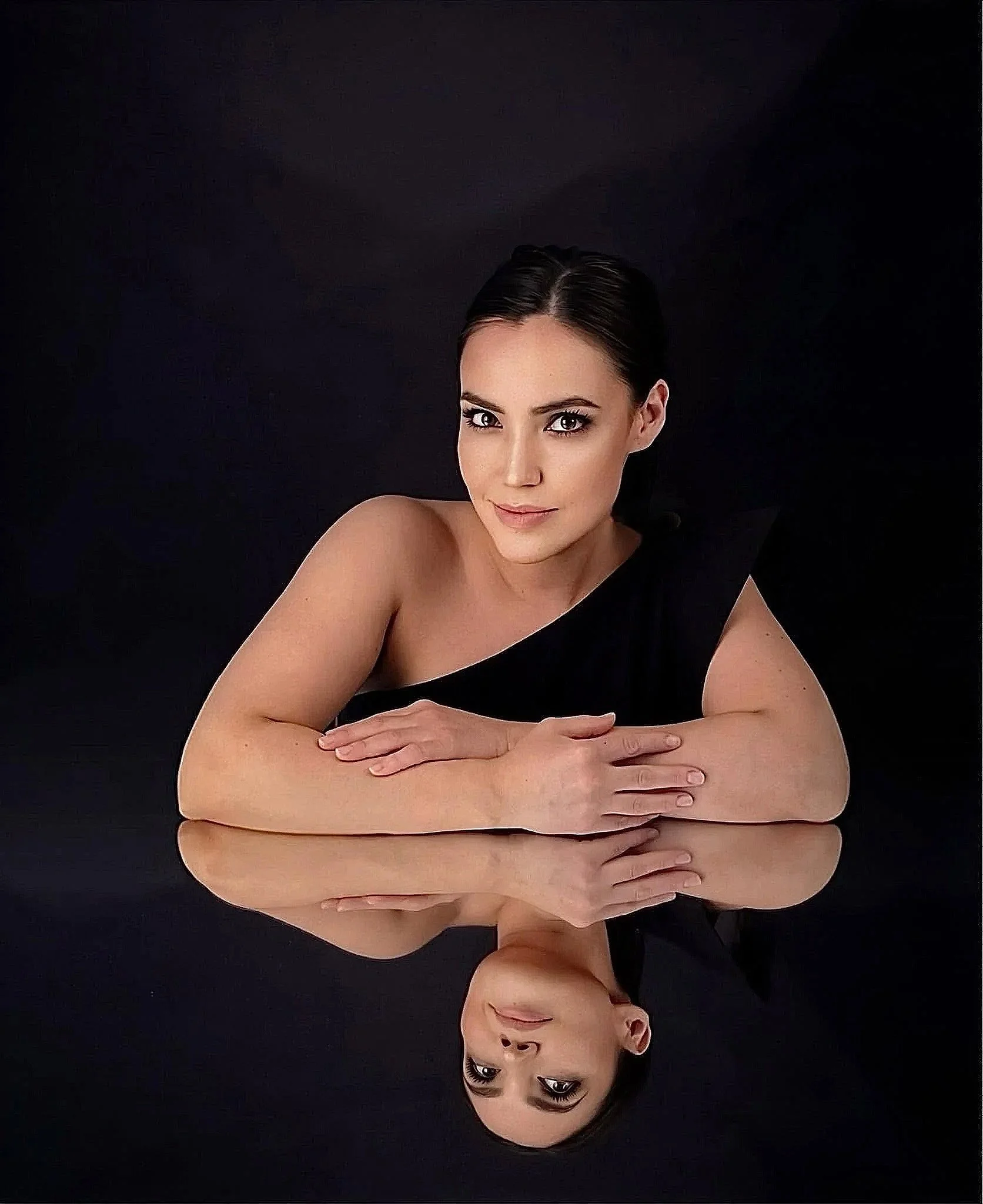Portrait of a woman with dark hair in a bun, wearing makeup, and posing with her arms crossed in front of her on a black background.