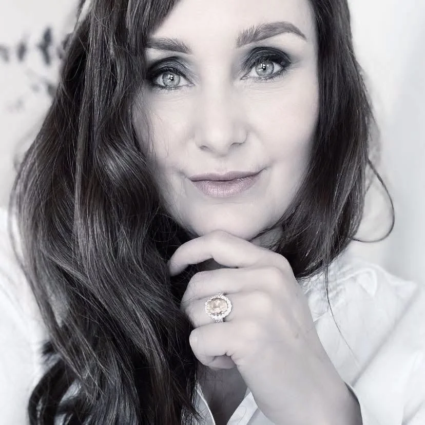 Close-up portrait of a woman with long dark hair and striking eyes, wearing a ring on her finger and touching her chin with her hand, in black-and-white.