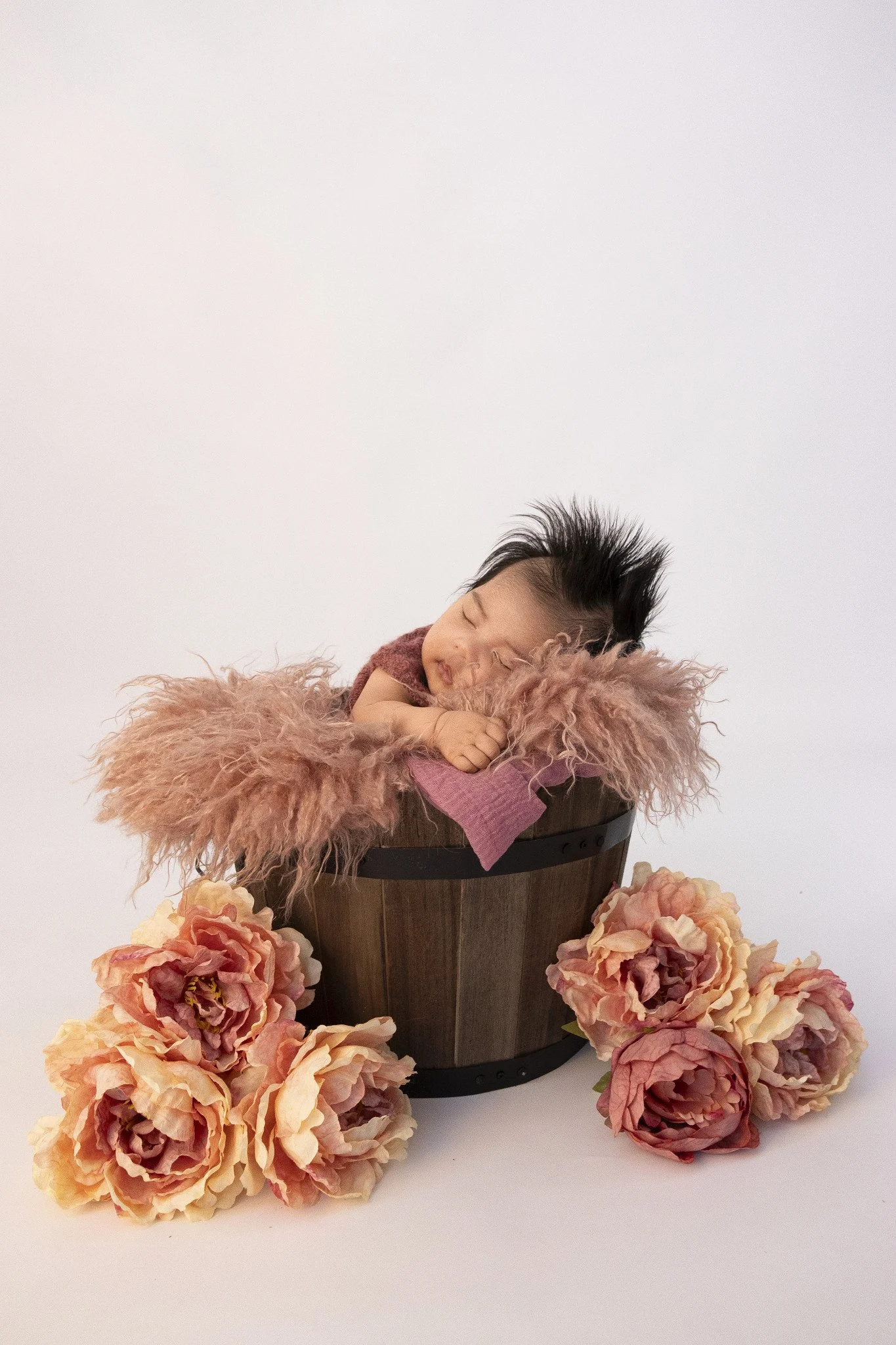 A sleeping baby with dark hair, resting on a fluffy pink blanket inside a wooden bucket, surrounded by pink and peach roses on a white background.