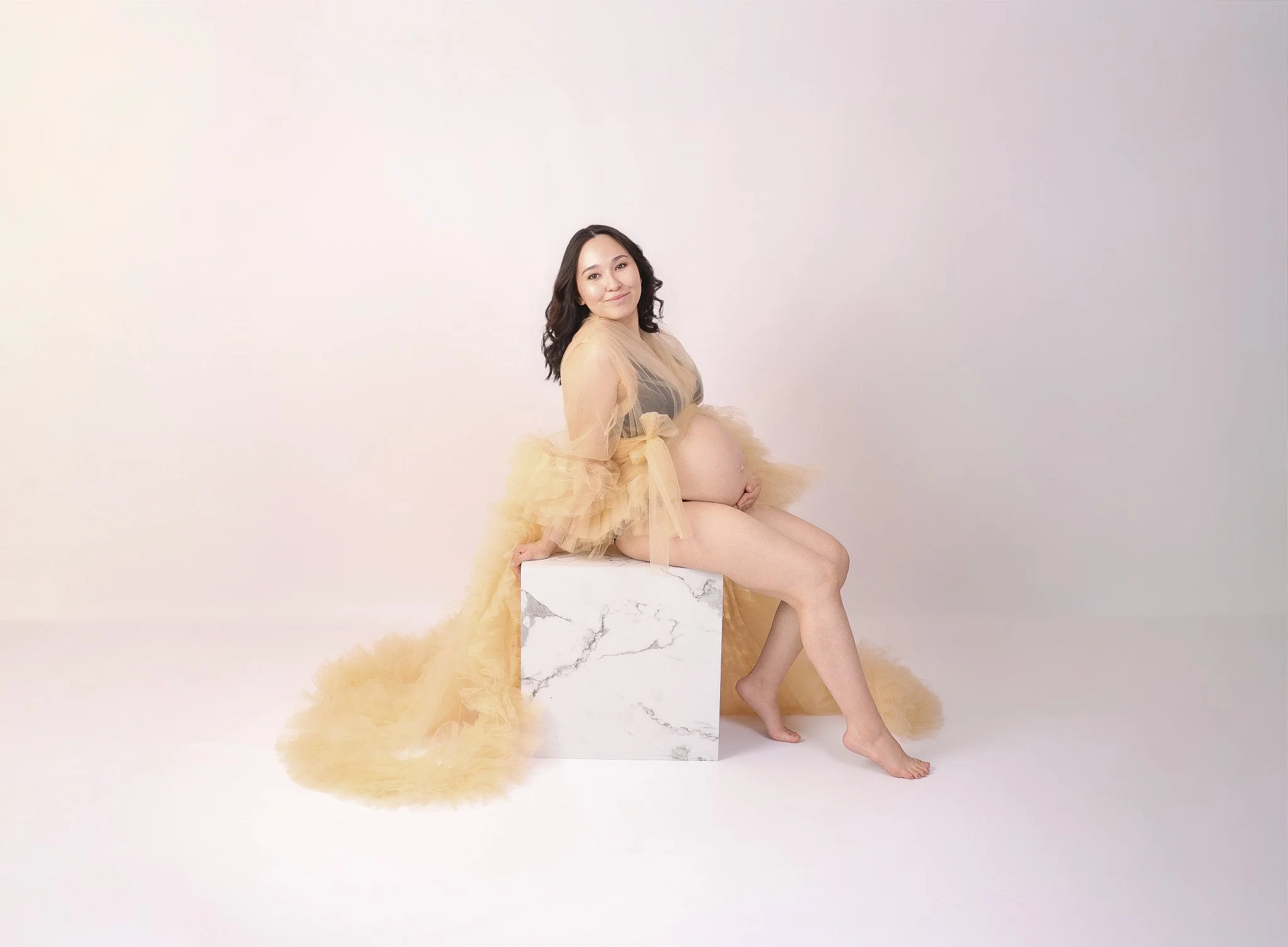 A pregnant woman wearing a beige tulle gown sitting on a marble pedestal, smiling at the camera, with a beige tulle train flowing onto the floor, against a plain off-white background.