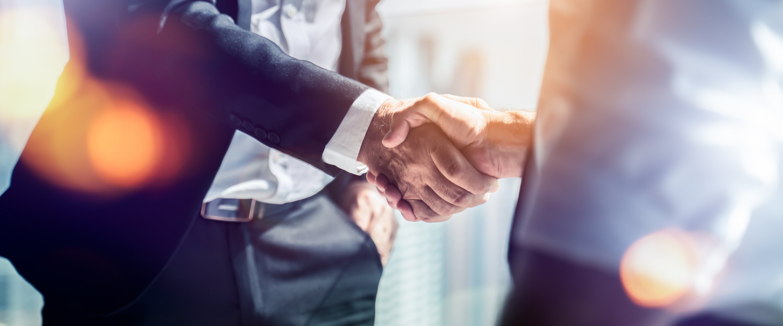 Close-up of two people shaking hands, one in a formal suit, in a business setting with bright lighting and a cityscape background.