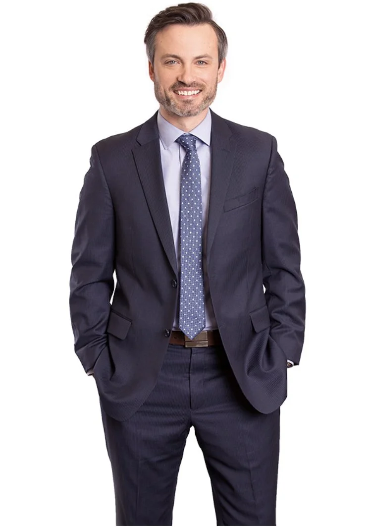 A man in a dark business suit with a light blue shirt and a blue patterned tie, smiling and standing with his hands in his pockets.